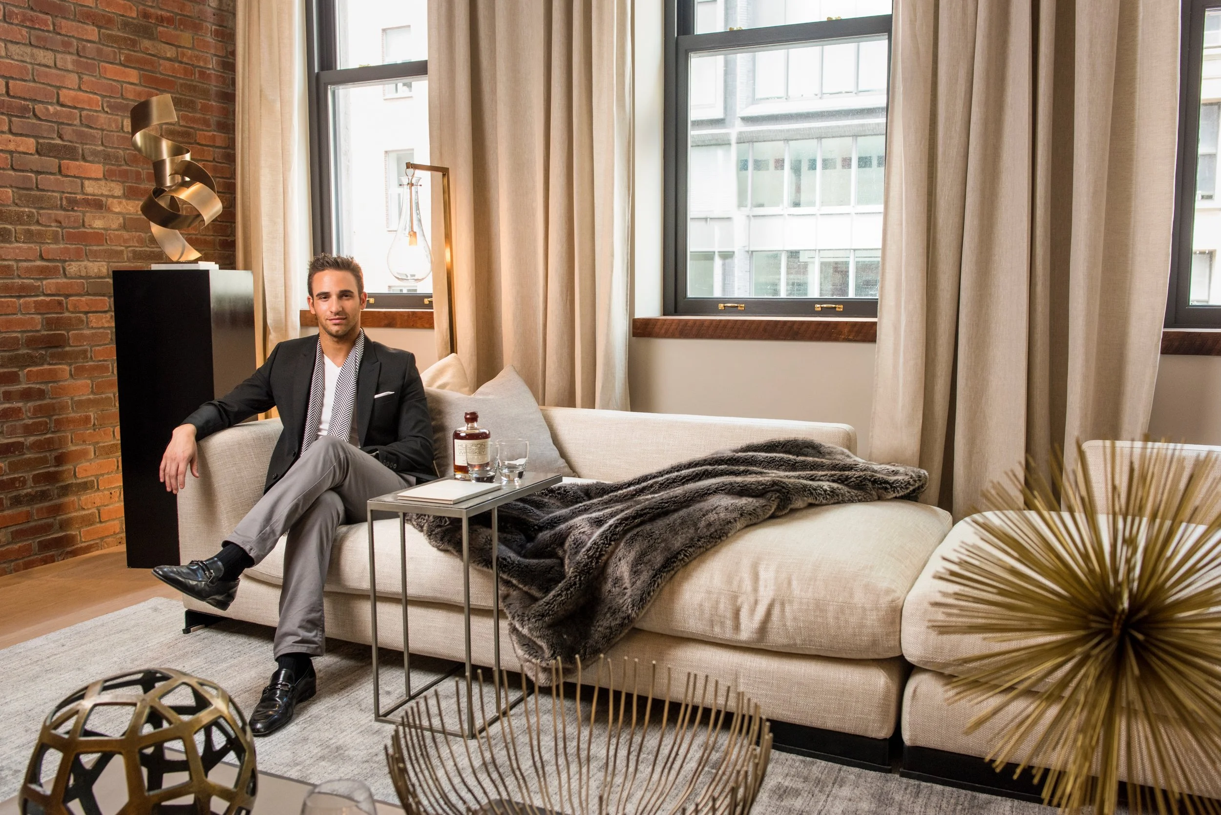A man in a black blazer, gray pants, and black shoes sitting on a beige sofa with a fur throw. There are large windows behind him with beige curtains, a small table with a bottle of liquor, a glass, and a notebook. The room has an exposed brick wall, decorative sculptures, and modern art pieces.