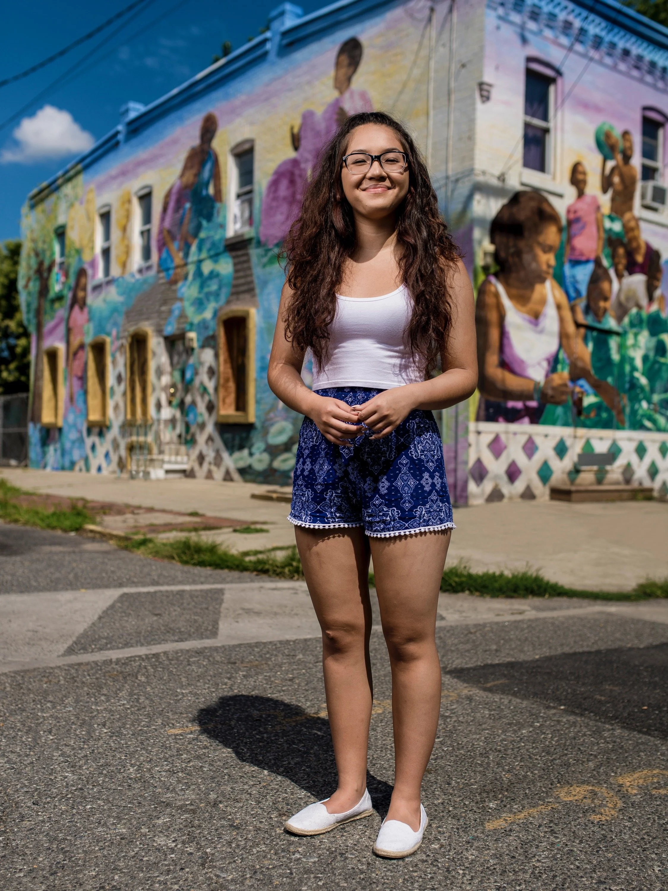 Young woman with glasses and long curly hair smiling in front of a colorful mural of diverse women painted on a building.