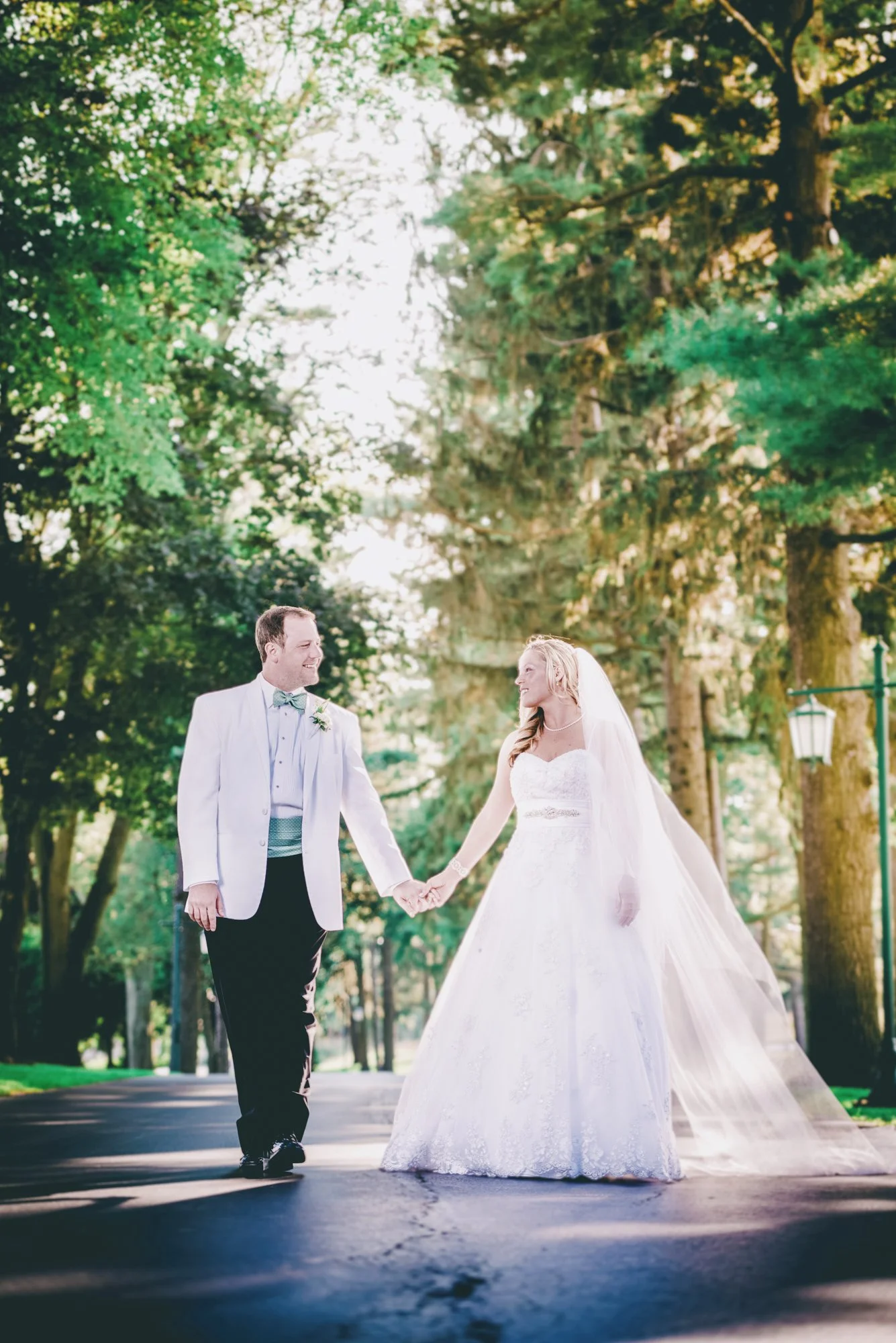 A newlywed couple holding hands and smiling while walking on a tree-lined path in a park, dressed in wedding attire with the bride in a white gown and veil, and the groom in a suit with a bow tie.