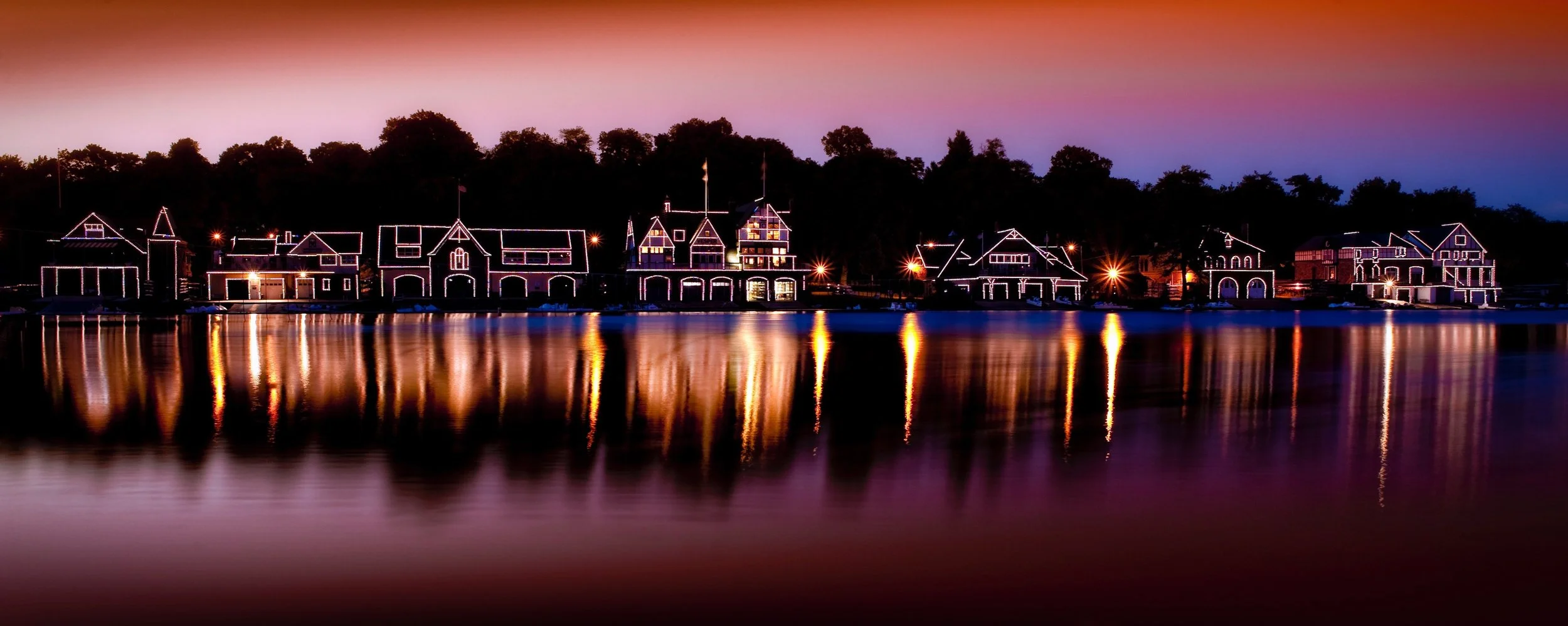 Nighttime view of houses along a waterfront decorated with string lights, their reflections visible in the water.
