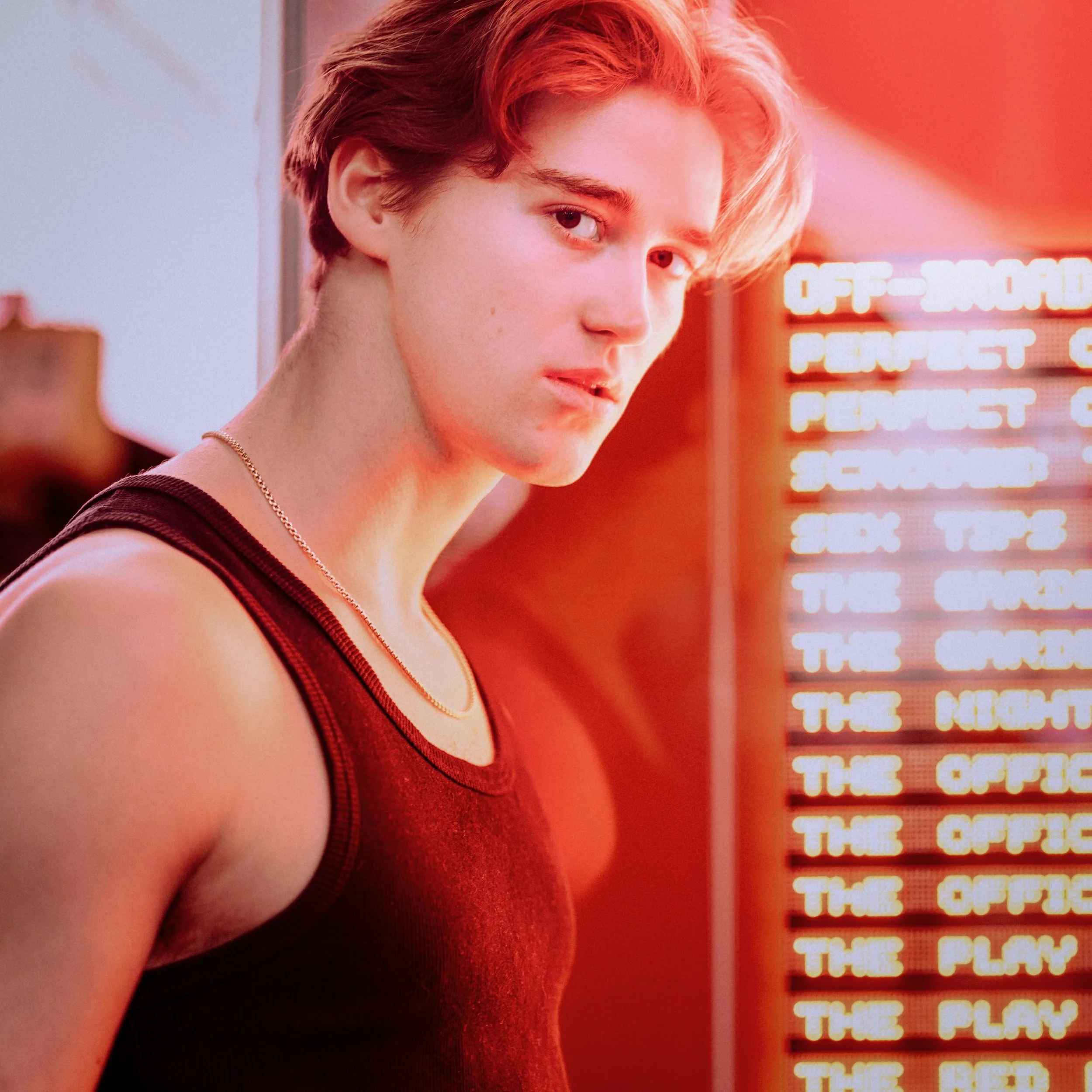 Young man with short hair wearing a sleeveless shirt and a chain necklace, looking over his shoulder in front of a neon sign.