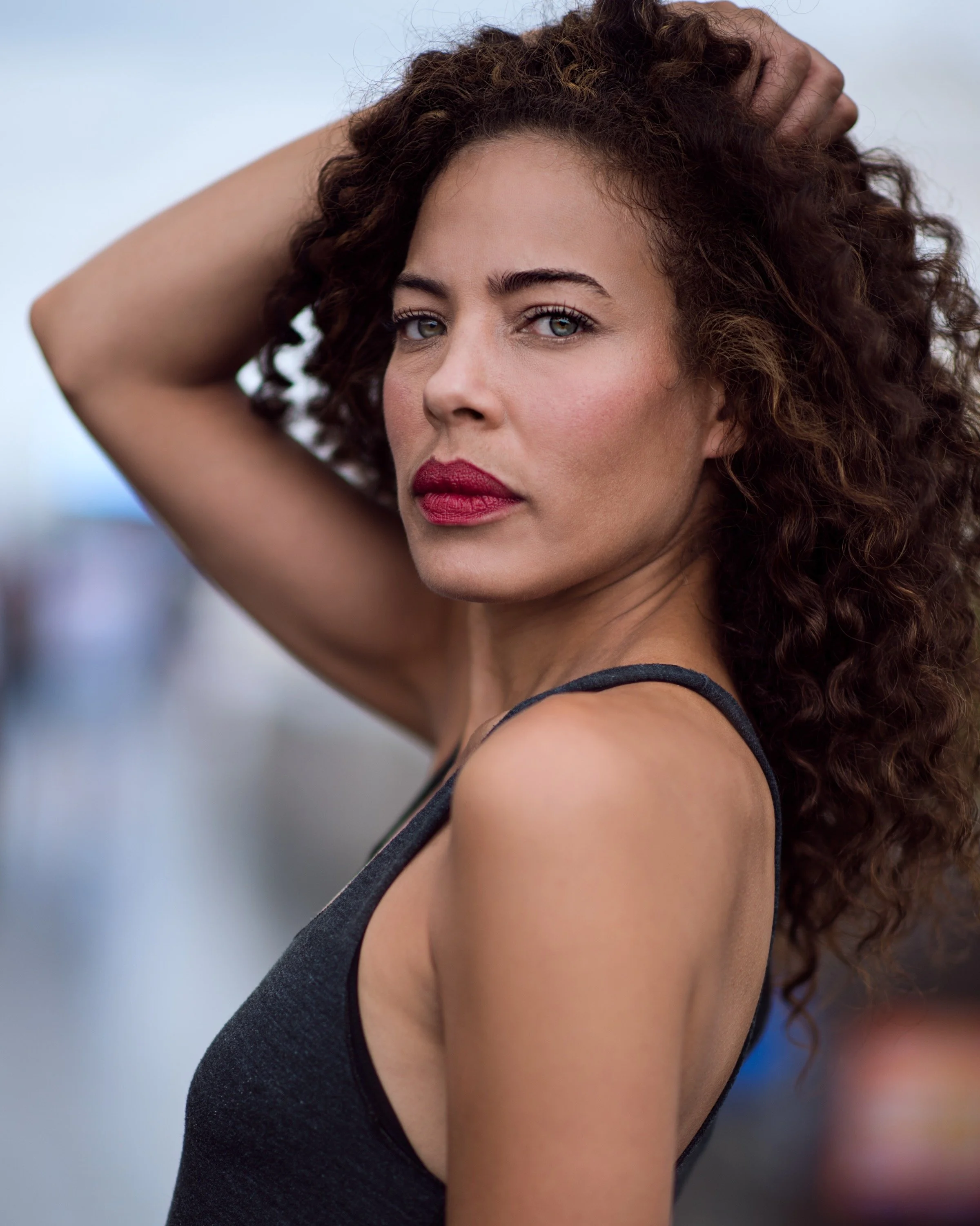 A woman with curly brown hair and fair skin wearing a black sleeveless top, posing outdoors with one hand on her head, looking directly at the camera.
