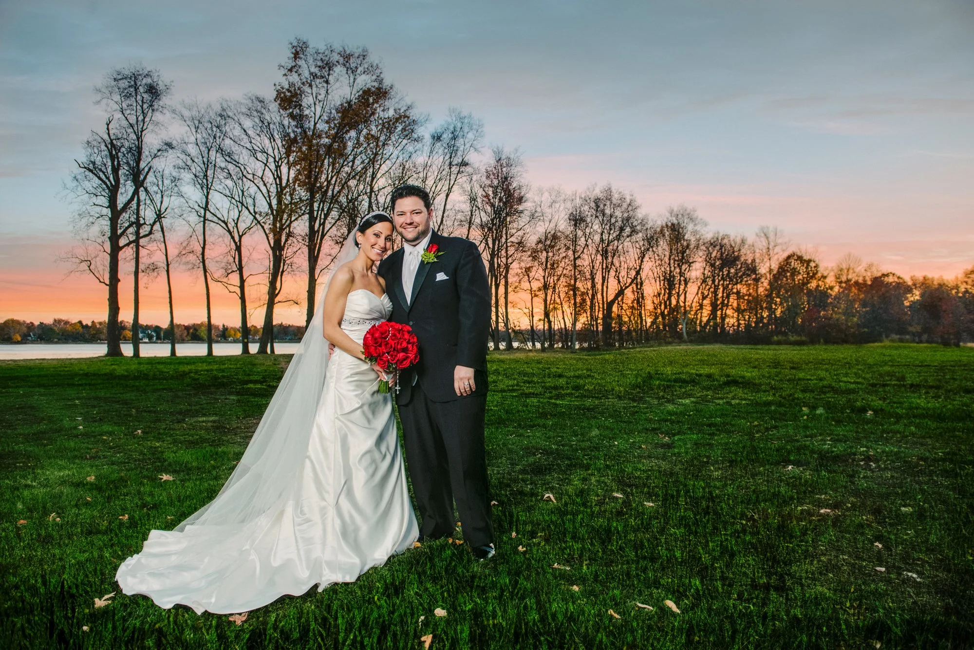 A newlywed couple standing on grass during sunset, with leafless trees and a lake in the background. The bride is wearing a white strapless wedding gown and holding a bouquet of red roses, while the groom is dressed in a black tuxedo with a red bouto