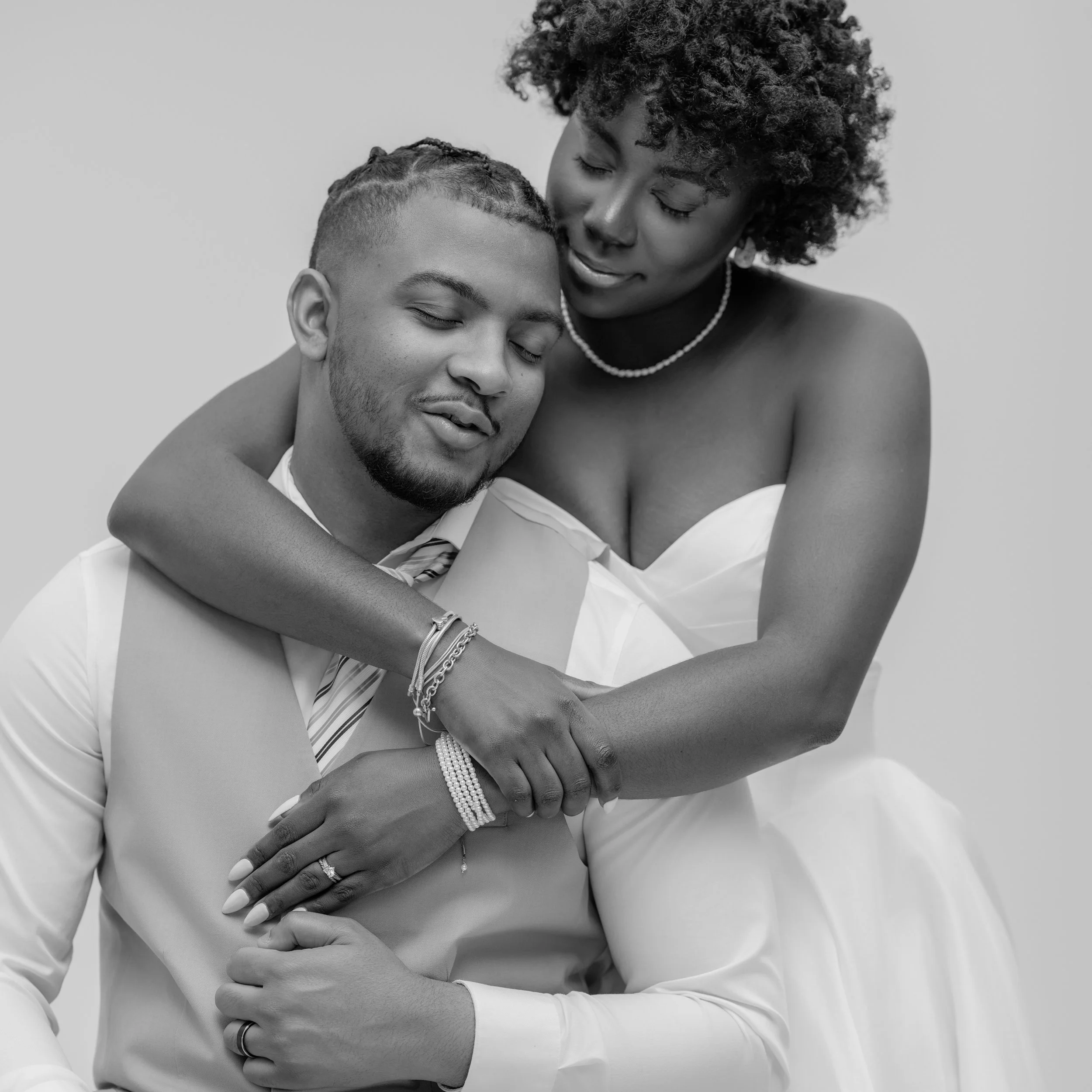 A black and white photo of a woman embracing a man. Both have their eyes closed and appear to be happy. The woman is wearing a strapless dress and pearl necklace, while the man is in a dress shirt and tie, with jewelry on his wrist and hand.