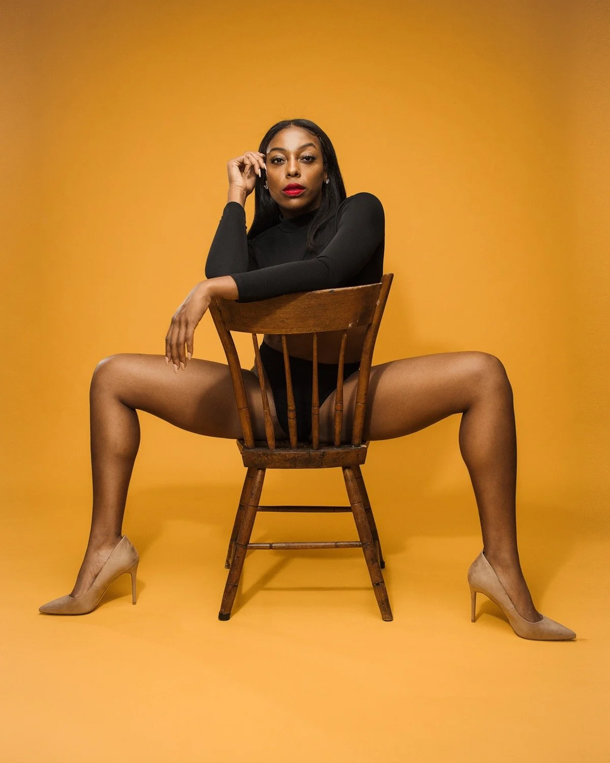 A woman sitting on a wooden chair with her legs spread apart, wearing a black long-sleeve top, black underwear, nude high heels, against a yellow background.