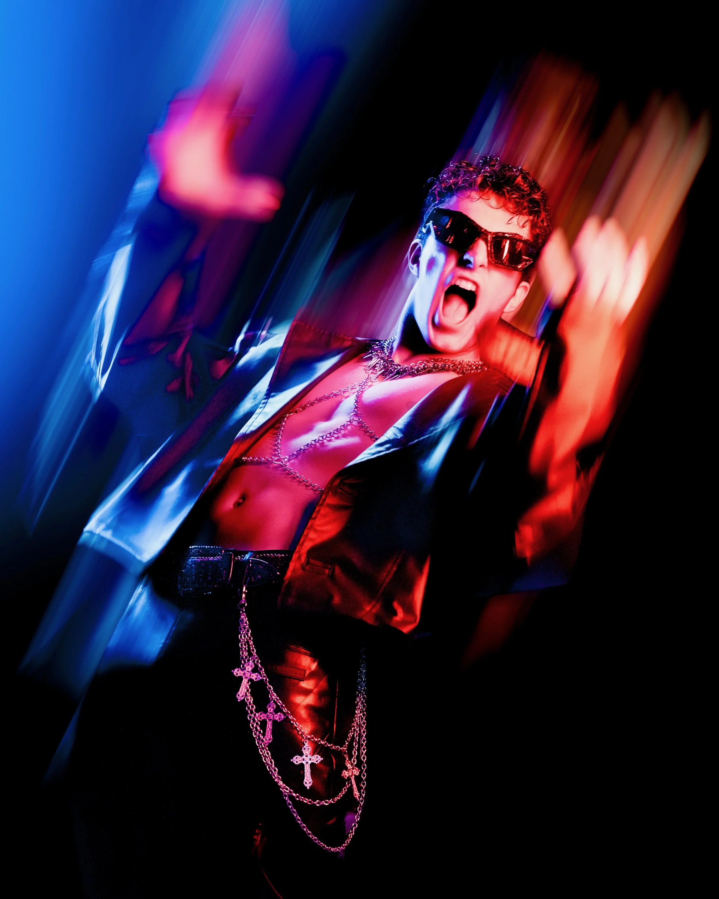 A person wearing sunglasses, necklaces, and chains, posing with an expressive, open-mouthed gesture against a colorful, blurred background.