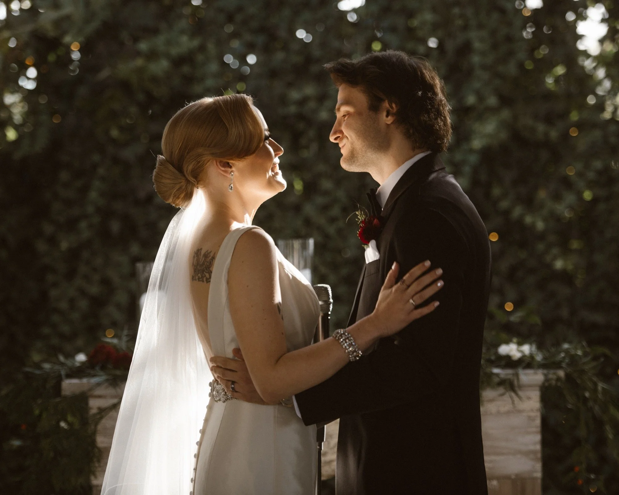 A bride and groom are standing close, holding hands, during their wedding ceremony outdoors, smiling at each other with trees in the background.