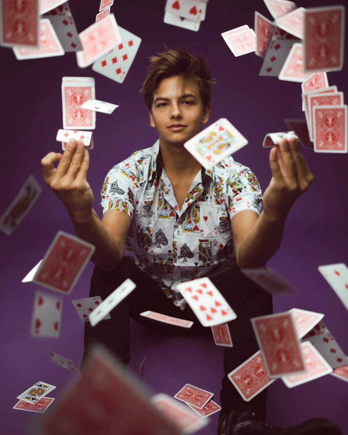 A young man with styled hair is sitting against a purple background, wearing a playing card patterned shirt, and lookings directly at the camera while playing with a large number of flying playing cards.