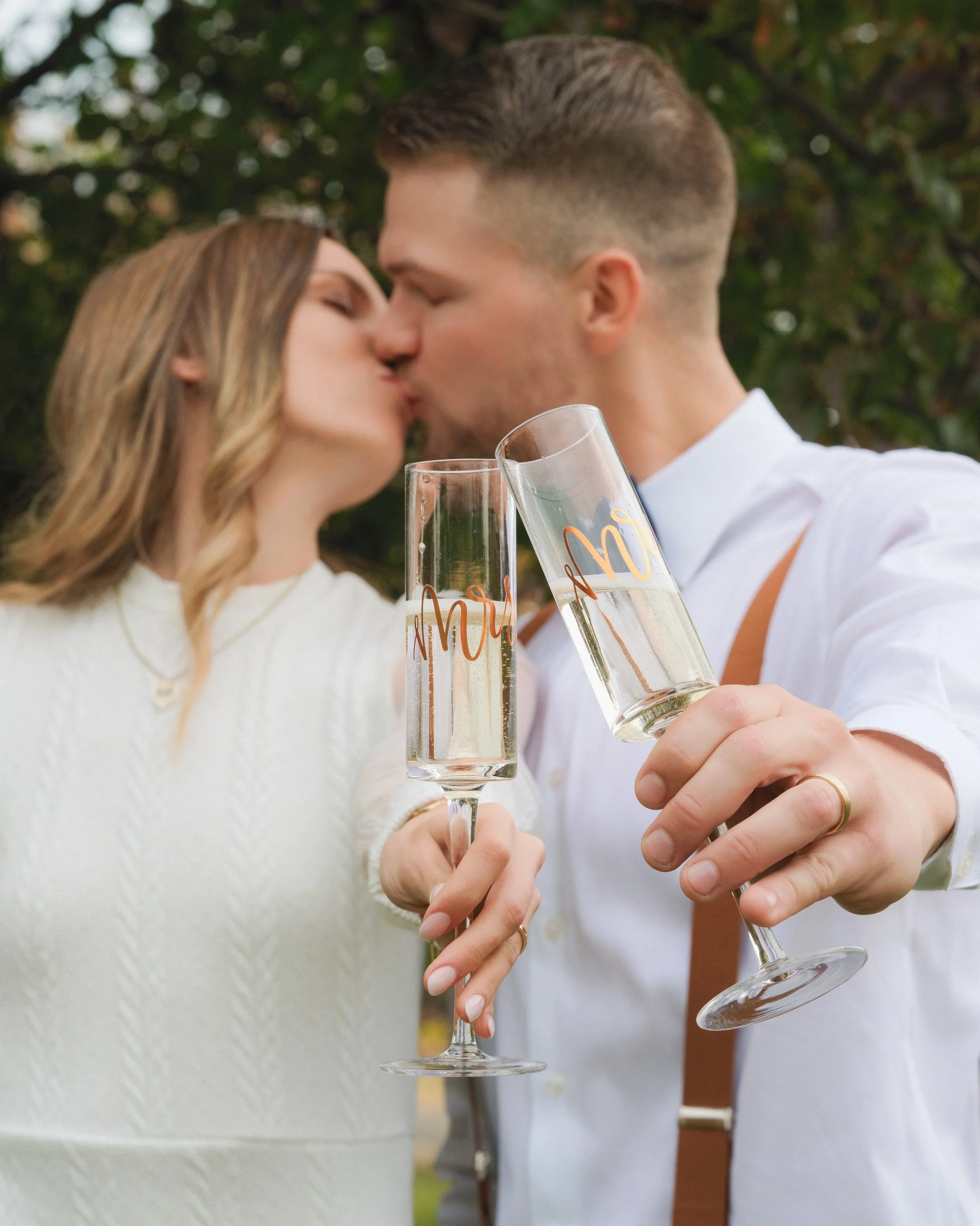 A couple is kissing outdoors while holding champagne glasses with 'Mr' and 'Mrs' written on them, celebrating their wedding.