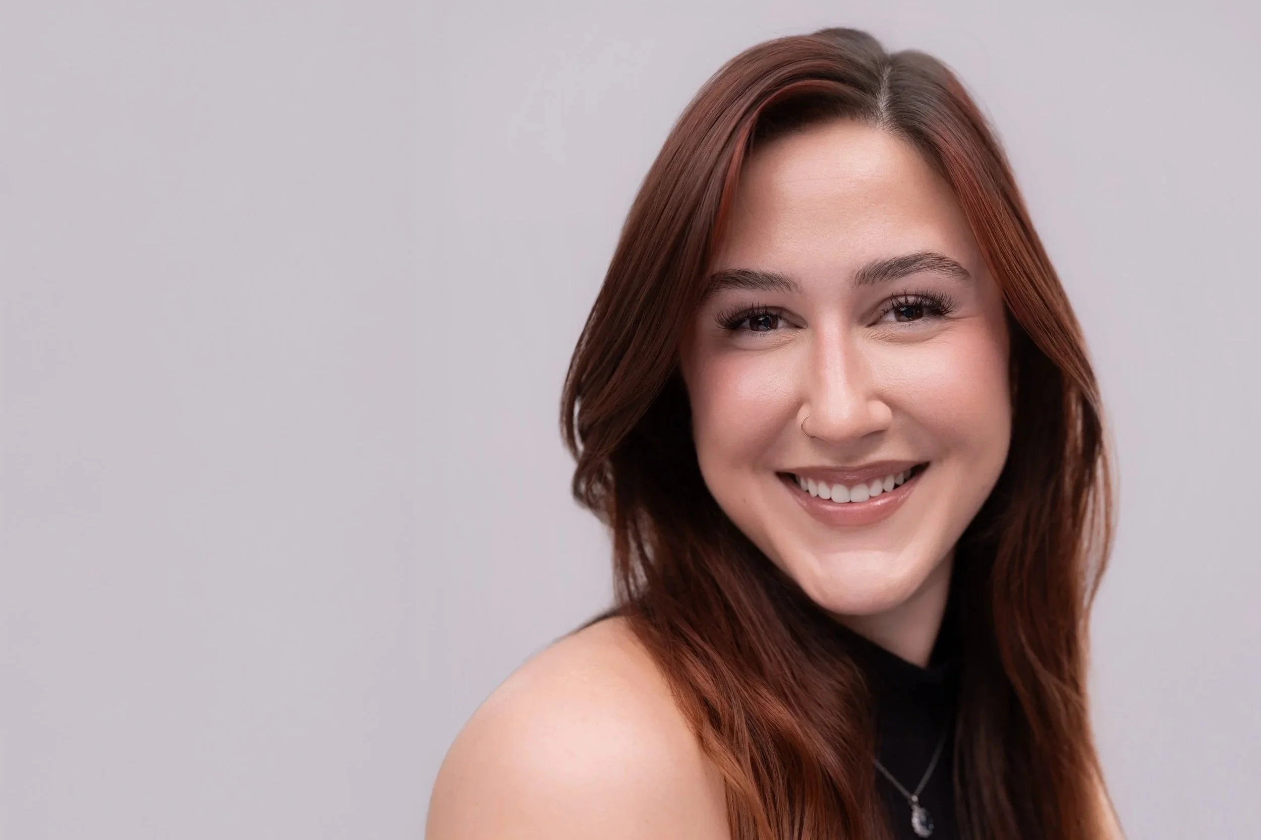 A young woman with shoulder-length auburn hair and a nose piercing smiling at the camera. She is wearing a black top and a necklace, standing against a plain light gray background.