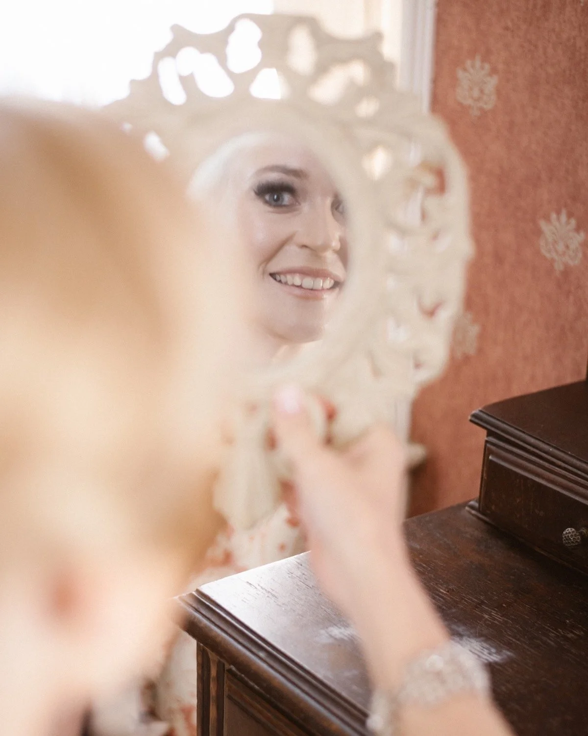 A young woman smiling at her reflection in an ornate hand mirror, sitting at a wooden table with a warm, patterned wall in the background.