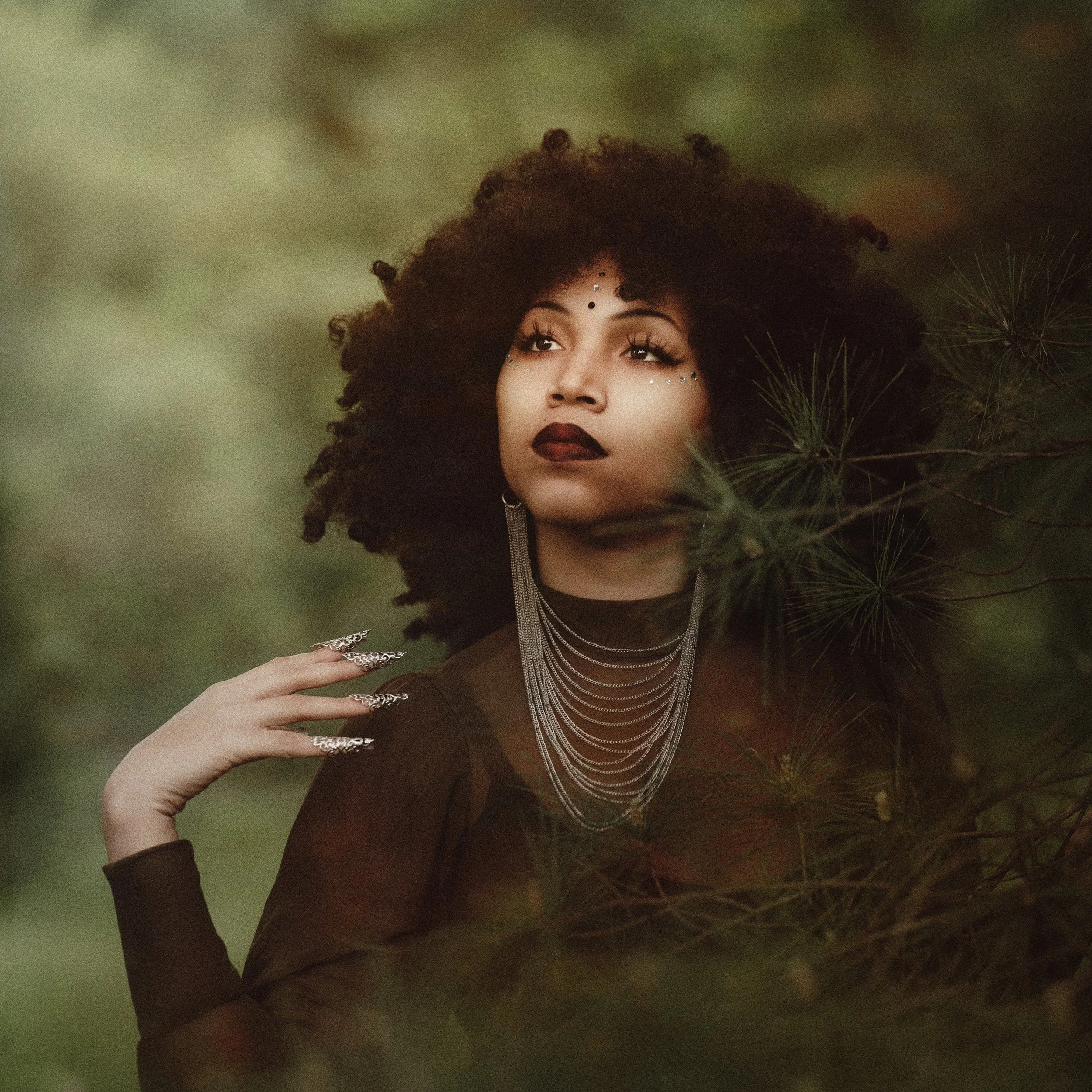 A woman with dark curly hair and dark lipstick, wearing layered necklaces and decorative face gems, looking contemplative amidst green foliage.