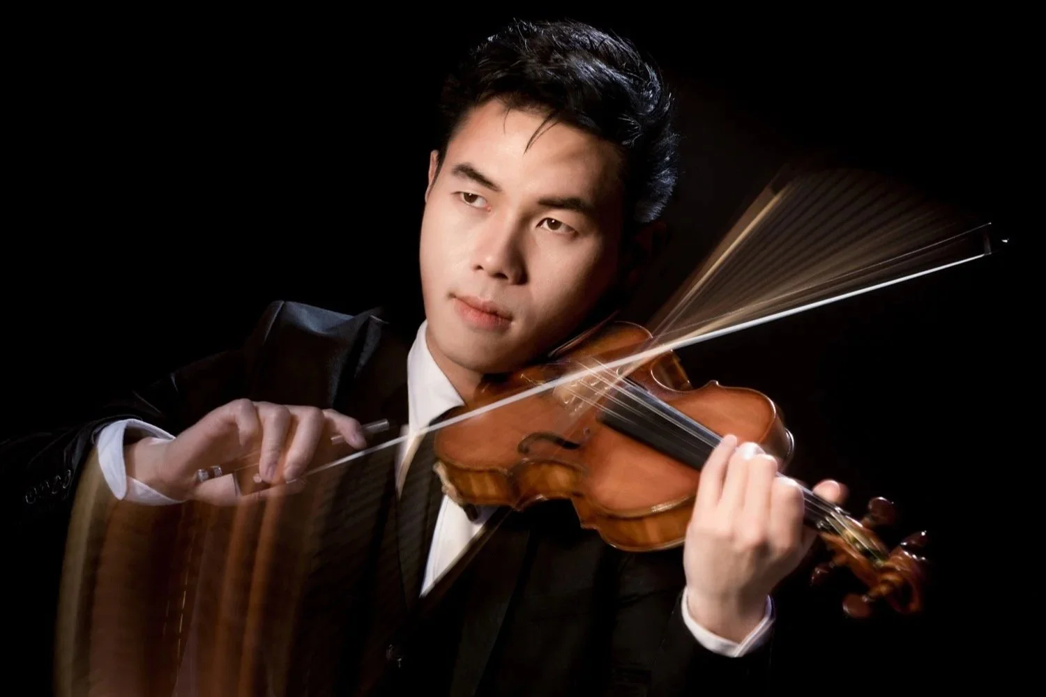 A man in a tuxedo playing a violin against a black background.