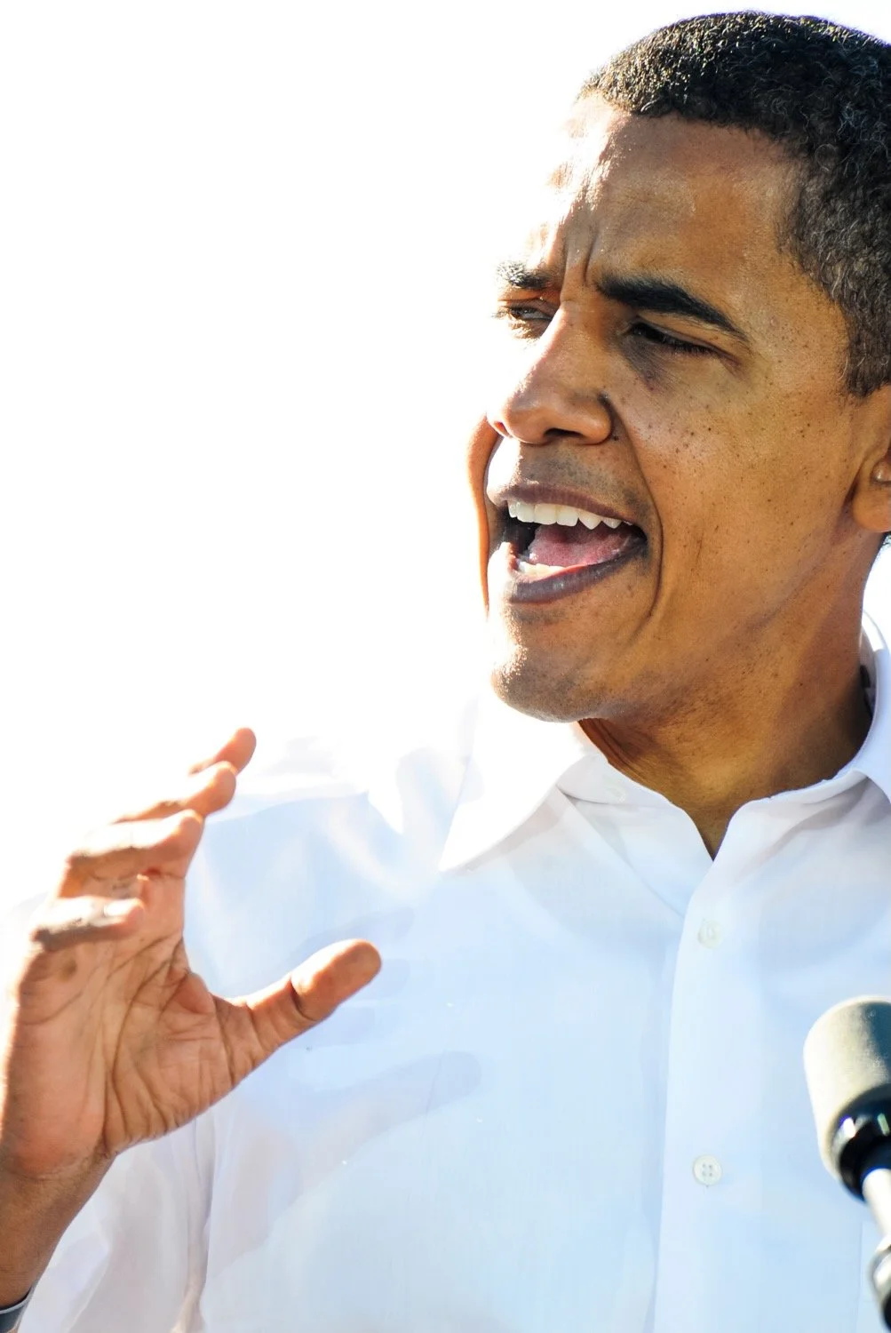Close-up of a man speaking passionately with a microphone, wearing a white shirt, against a bright light background.