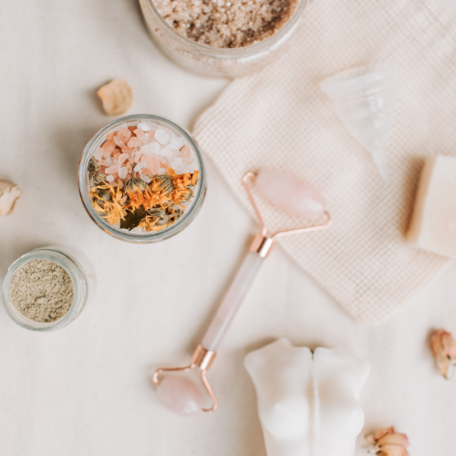 Beauty tools and supplies on a white surface, including a facial roller with pink stones, dried flower bath salts, a jar of bath salts, a small container of loose powder, a soap bar, and a white cloth.