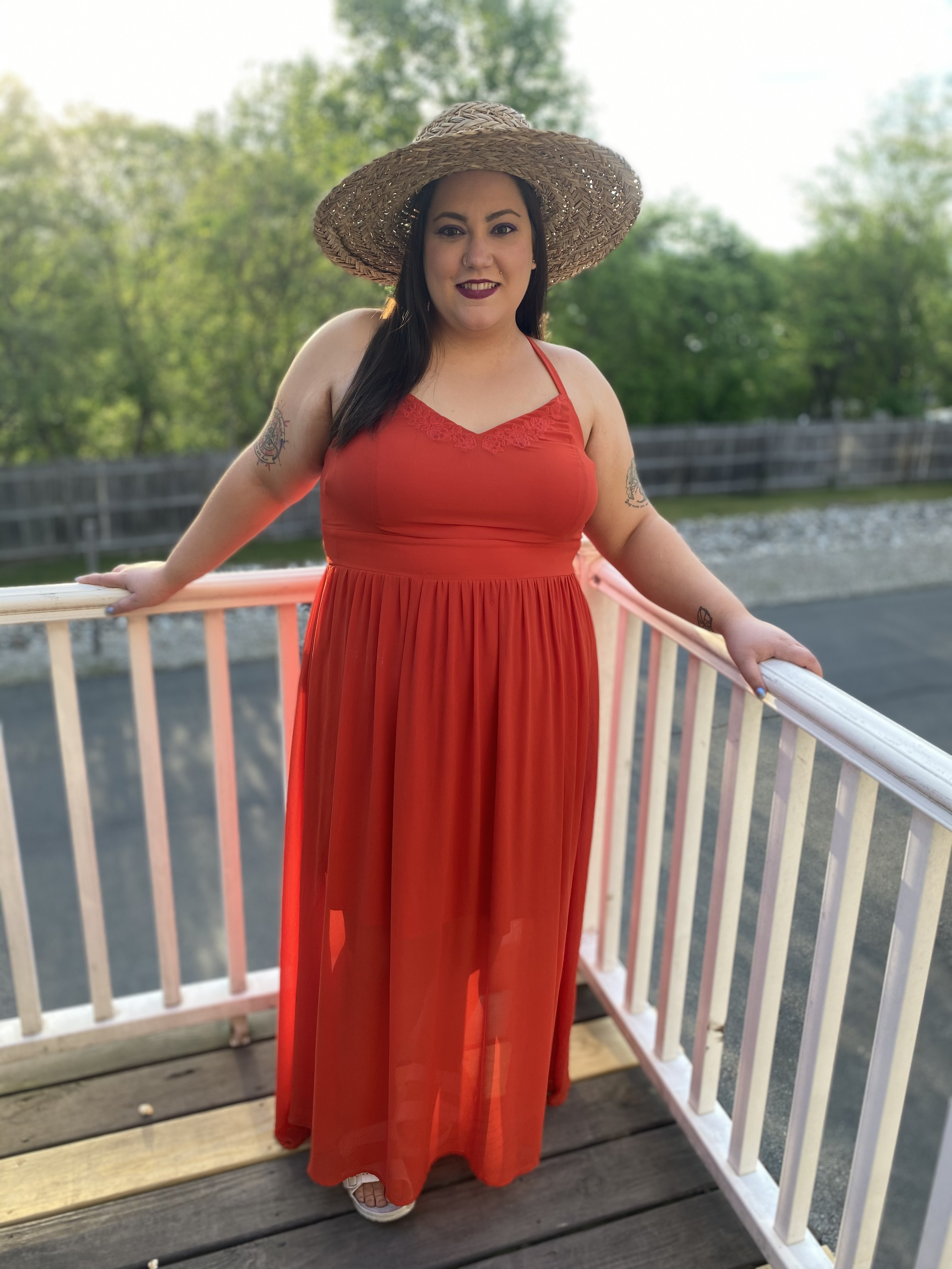 Amy, a dark-haired, brown-eyed, tattooed, fat white woman, stands leaning against a balcony with a large smile. She wears a long orange maxi dress and a large straw sun hat.