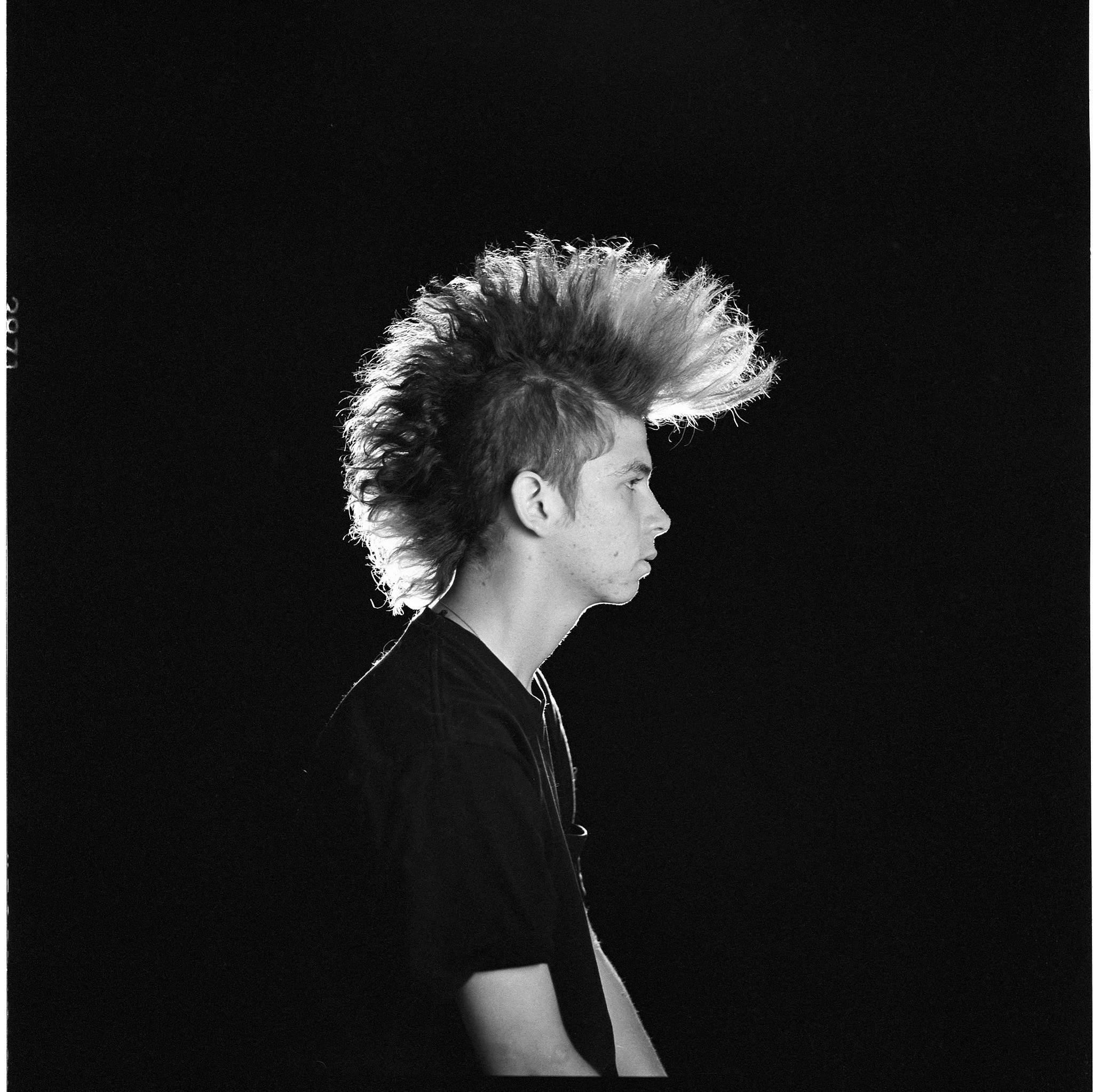 Profile of a young man with a mohawk hairstyle, facing right, in black and white against a dark background.