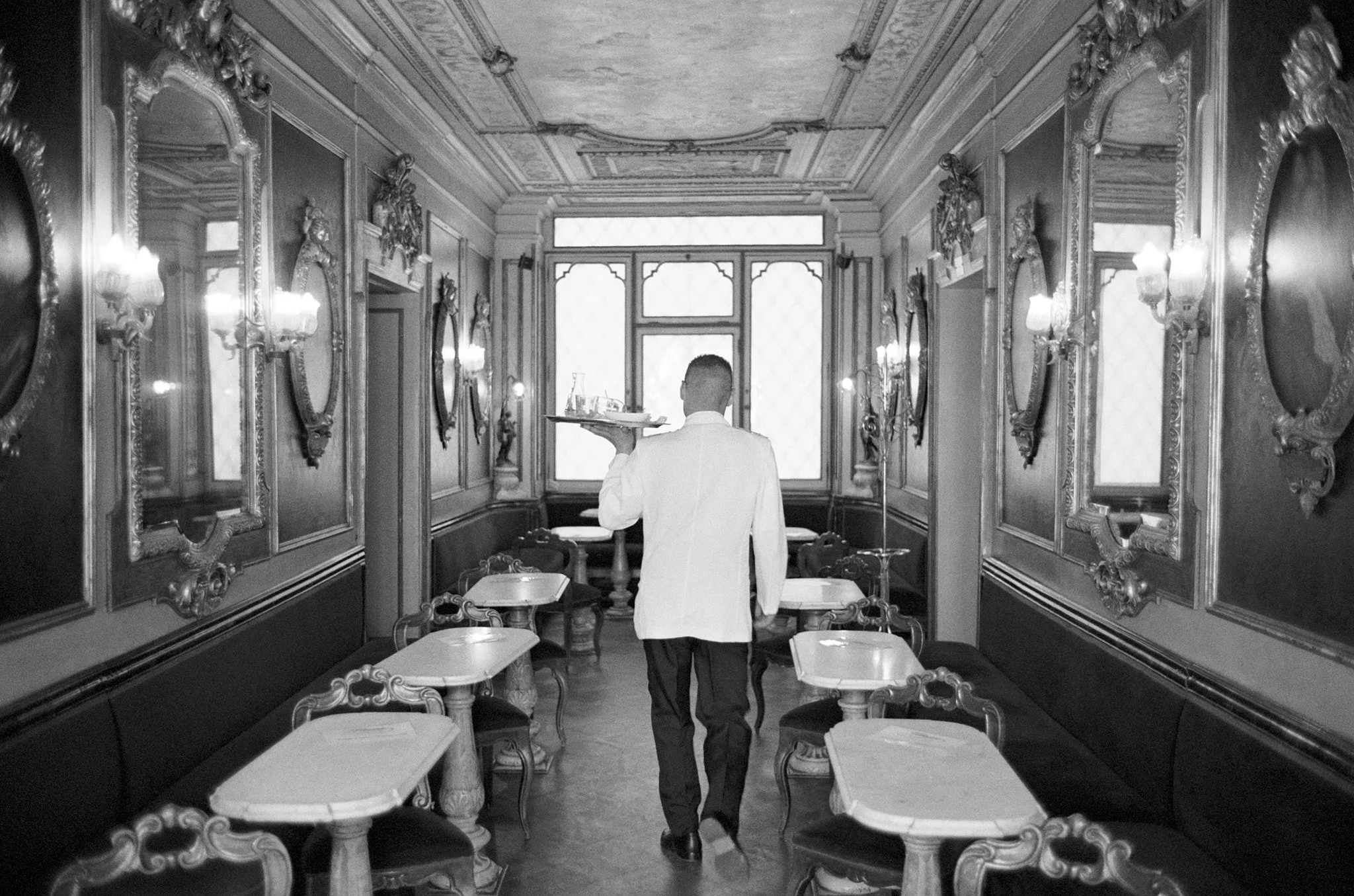 A waiter in a white coat serving drinks in an ornate, vintage-style restaurant with decorated walls, mirrors, and tables.