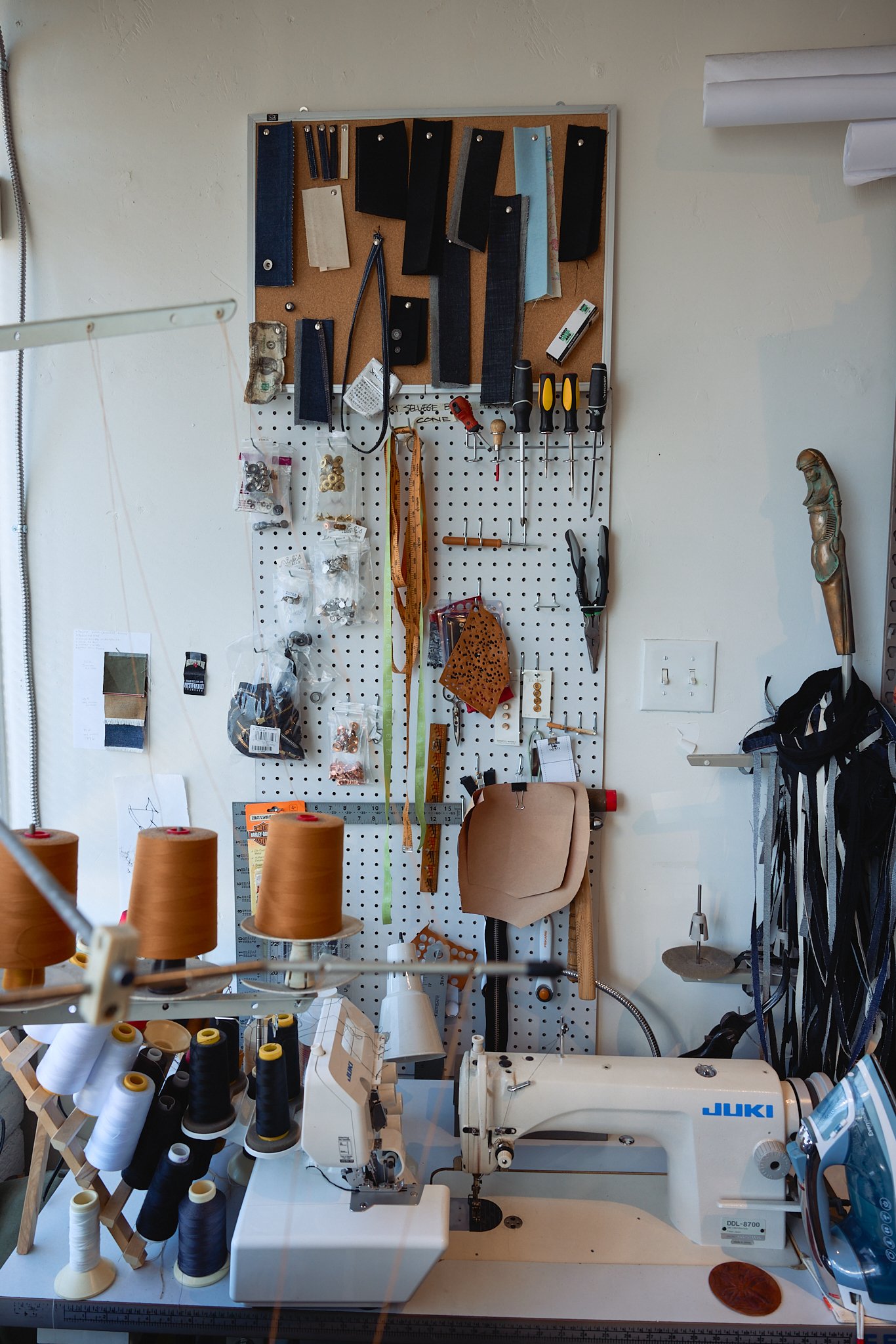 A sewing workspace with a JUKI sewing machine on a table and a pegboard wall holding tools, fabrics, threads, and sewing accessories.