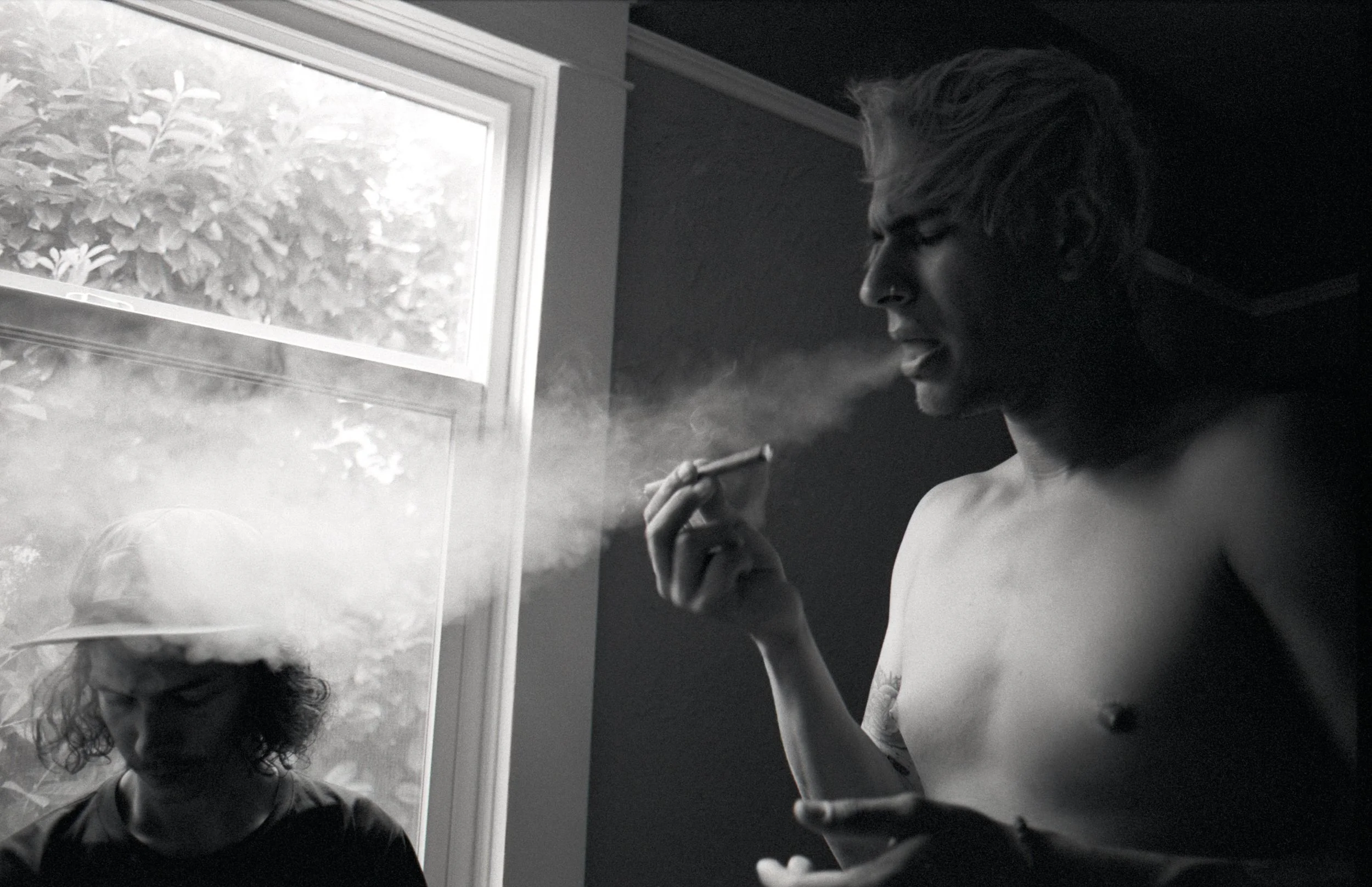 Black and white photo of a shirtless young man smoking a cigarette near a window, with another person with curly hair and a cap visible through the window.
