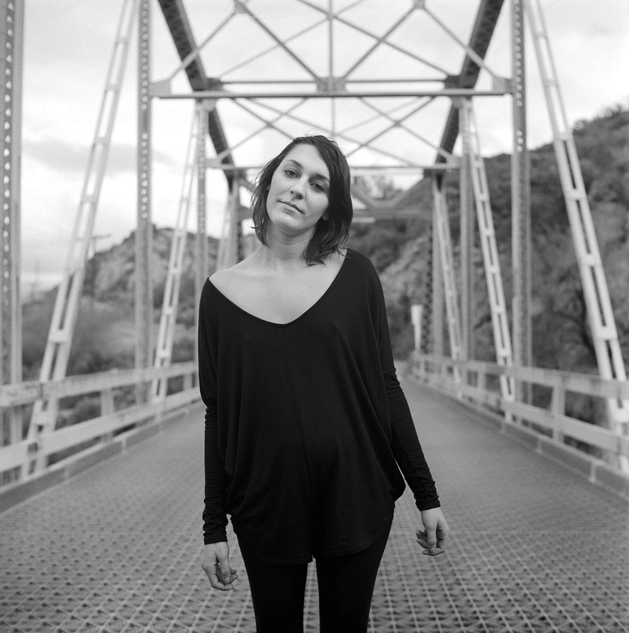A woman in a black long-sleeve shirt and pants standing on a bridge with a metal framework in a natural landscape.