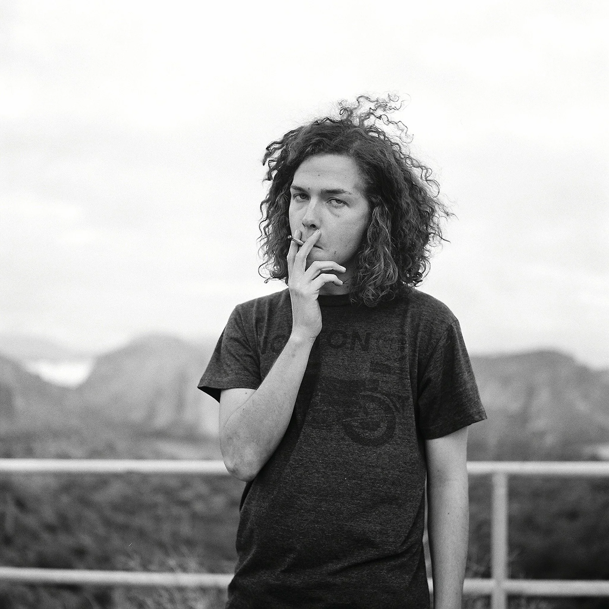 Black and white photo of a person with curly hair smoking a cigarette outdoors with mountains in the background.