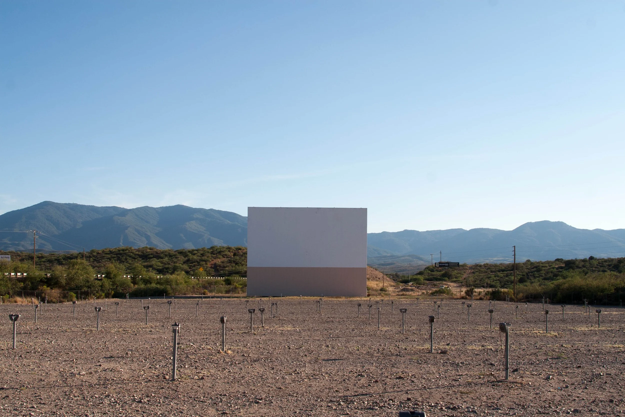 A large white and beige movie theater screen in a field with a mountain range in the background under a clear blue sky.
