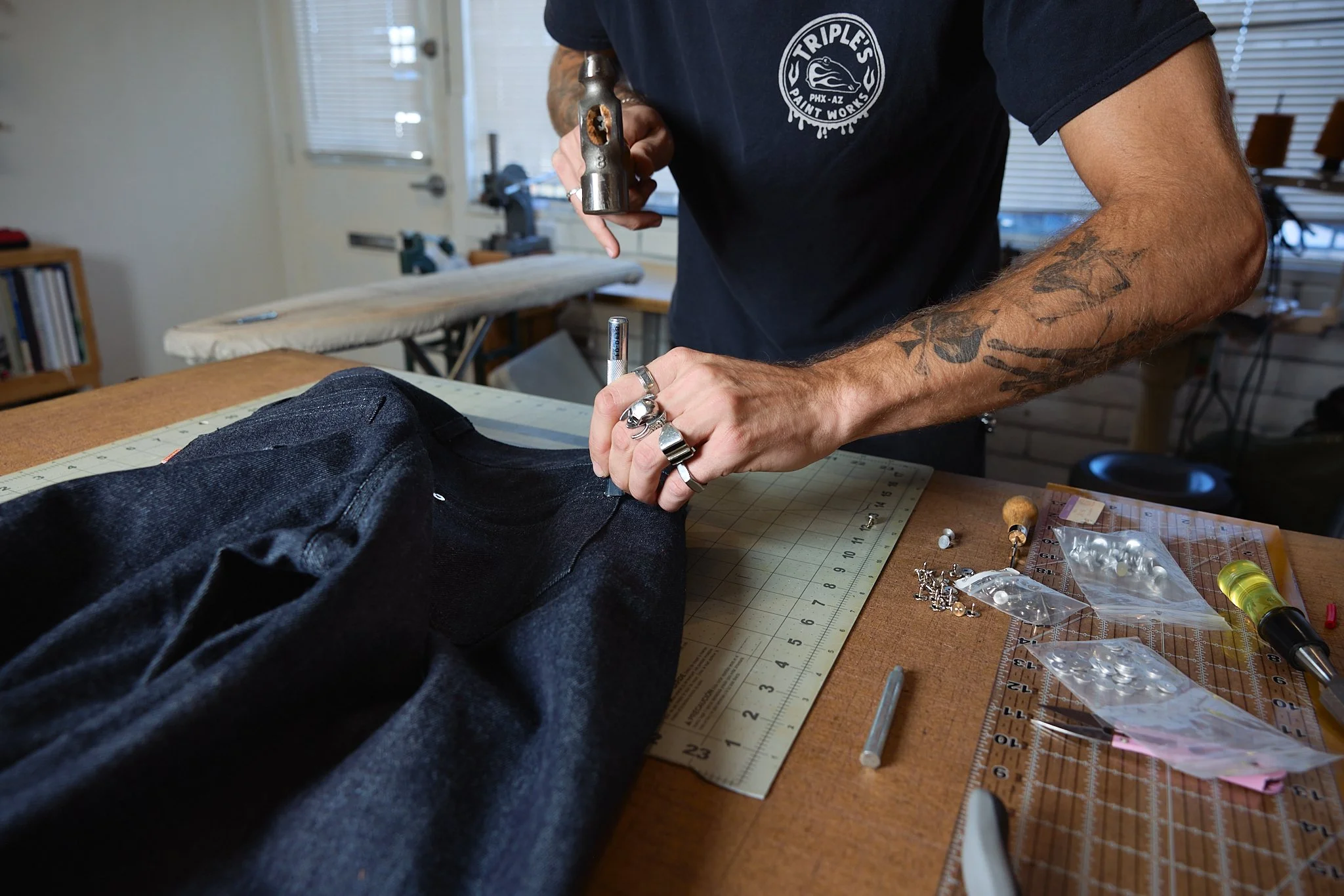 A person working on a pair of black jeans at a workbench with tools and materials around, including a hammer, screw, and packaging of buttons.