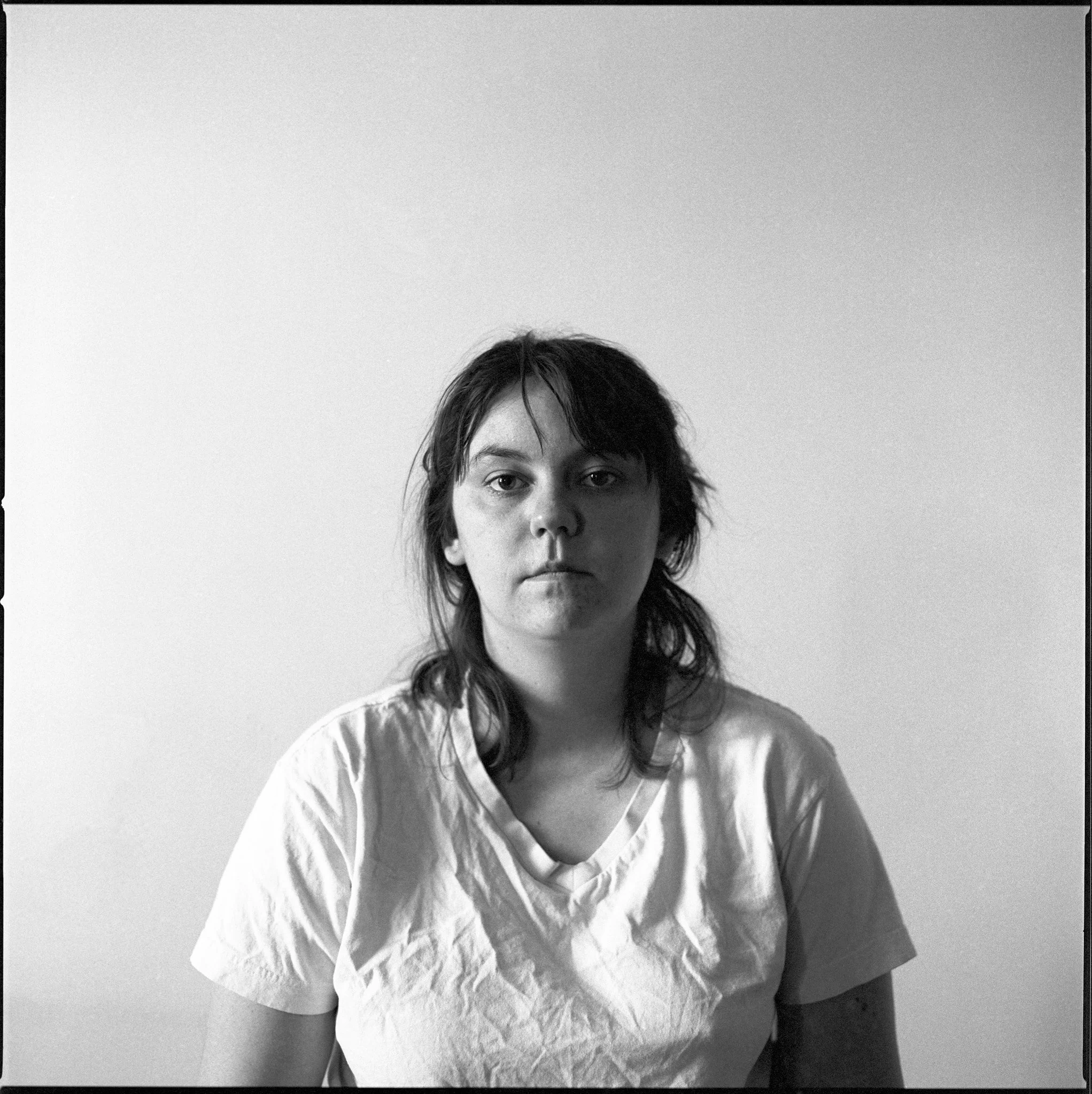 A black-and-white portrait of a woman with dark, messy hair, wearing a wrinkled T-shirt, looking directly at the camera with a neutral expression against a plain light-colored wall background.