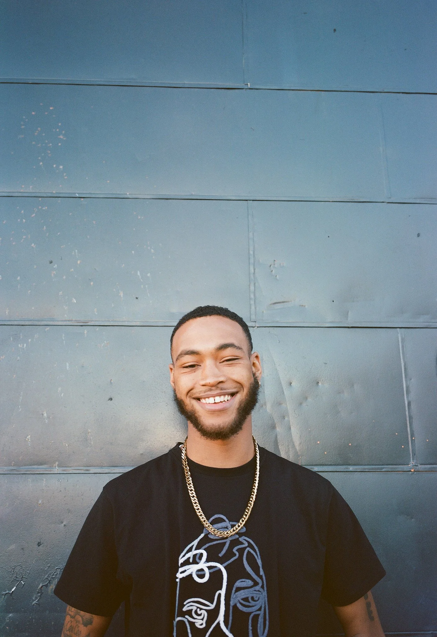 A smiling young man with a beard and short hair, wearing a black t-shirt with abstract line art, a gold chain, standing in front of a blue wall.