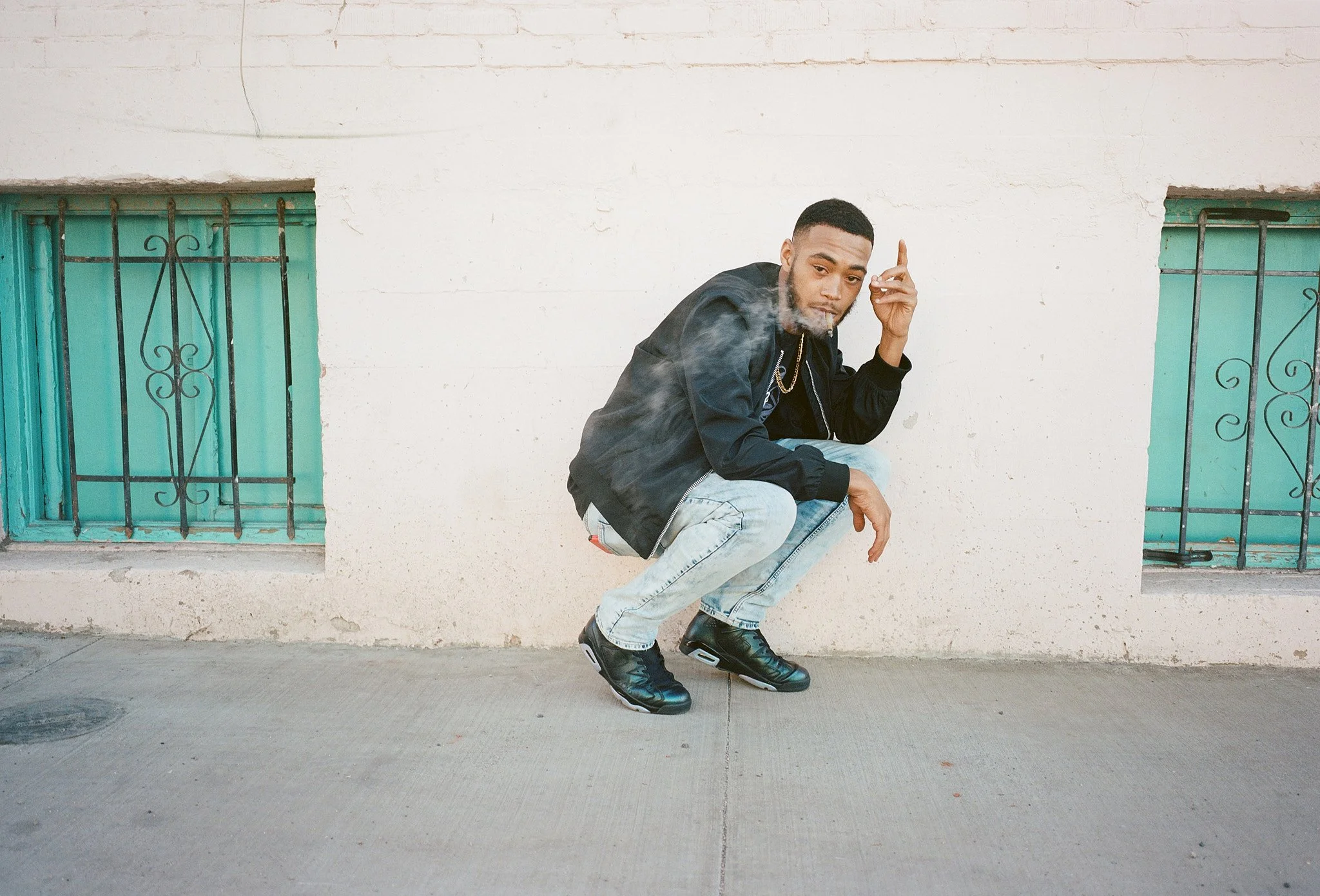 A young man squatting in front of a white brick wall with bars on windows, smoking and making a gesture with his hand.