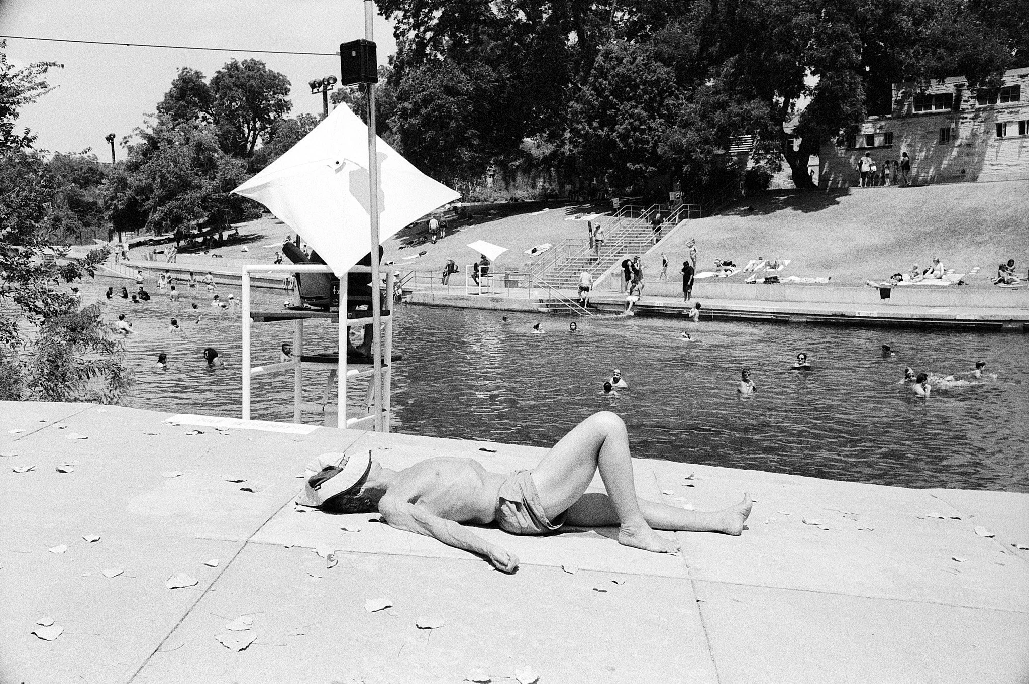 A person lying on a towel by a swimming pool, wearing a hat, with people swimming and relaxing around the pool and on the grass in the background.