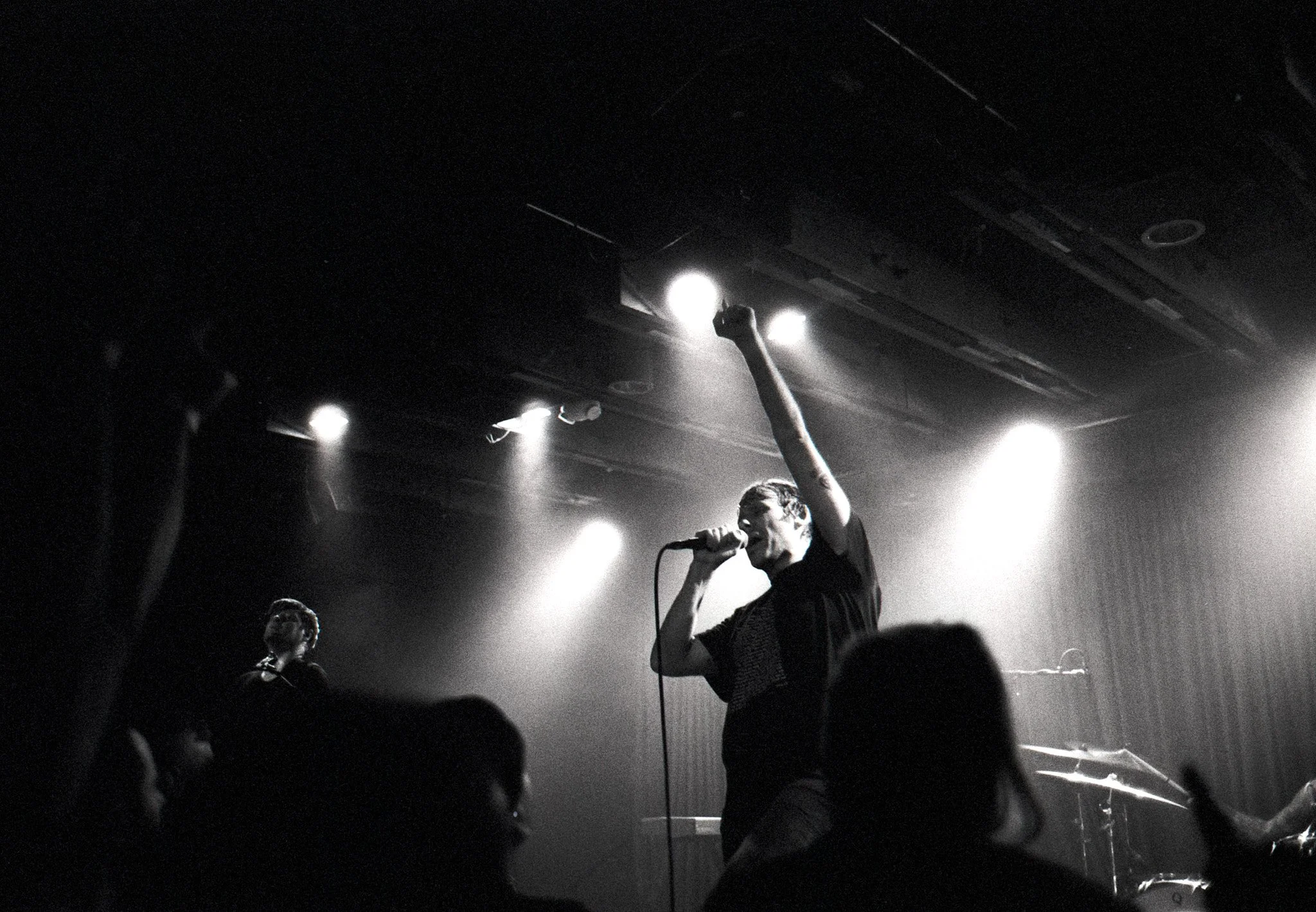 A black and white photo of a male singer on stage, holding a microphone with one hand raised, performing passionately during a concert. There are bright stage lights overhead and audience members visible in the foreground.