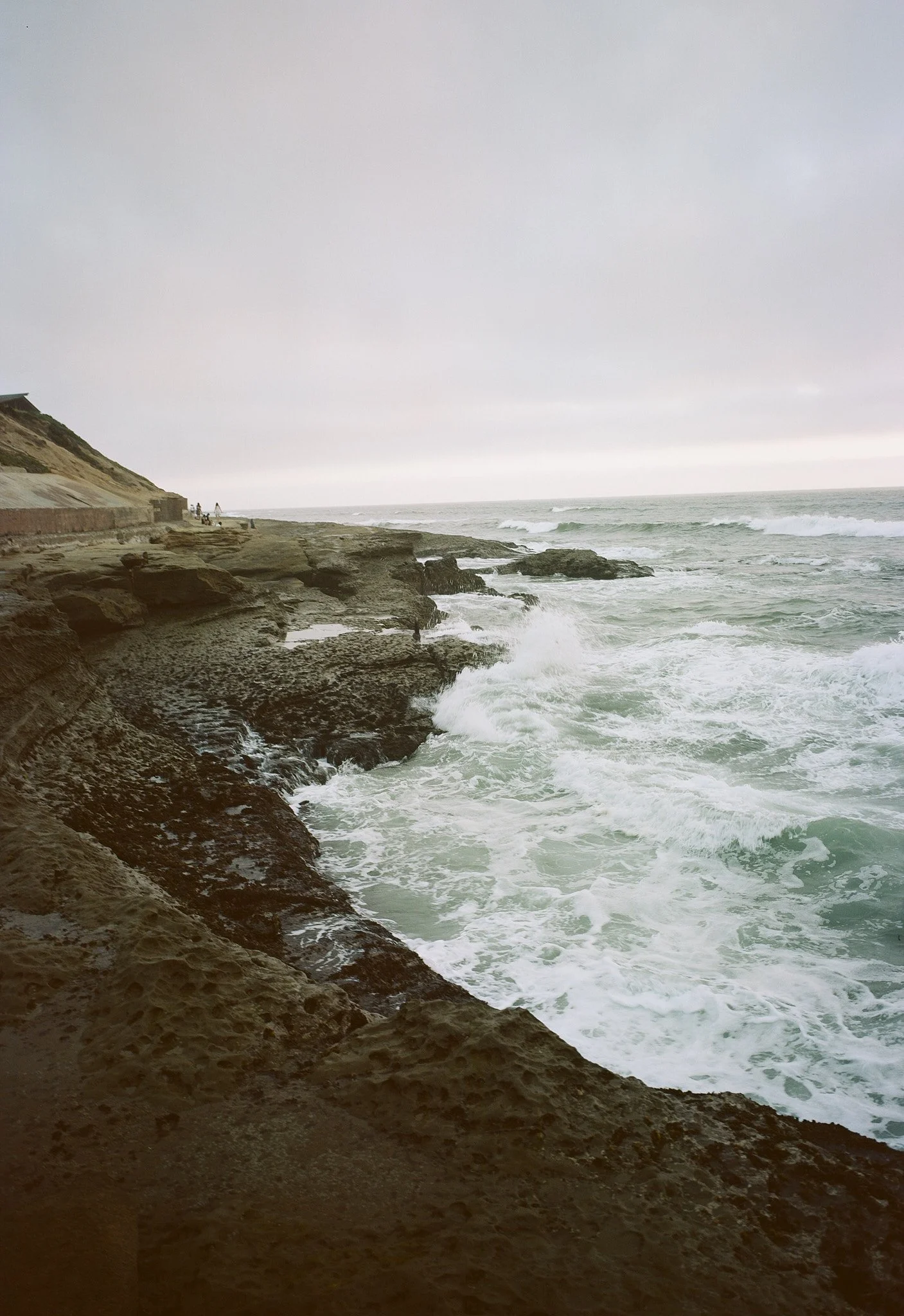 View of rocky coastline with waves crashing against the rocks during overcast day, with a cloudy sky and some people visible in the distance.