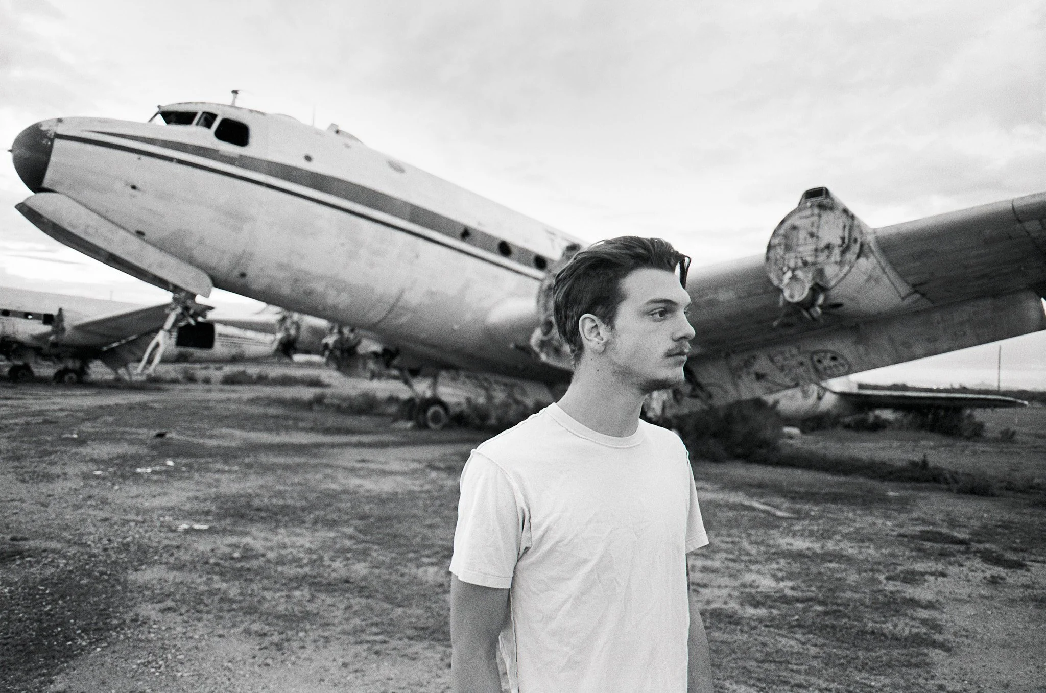 A young man in a white T-shirt stands outdoors near an abandoned boat and airplane on a barren landscape.