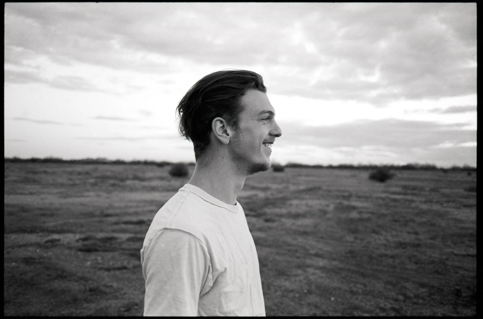 Side profile of a young man smiling outdoors in a wide open field with cloudy sky in black and white.