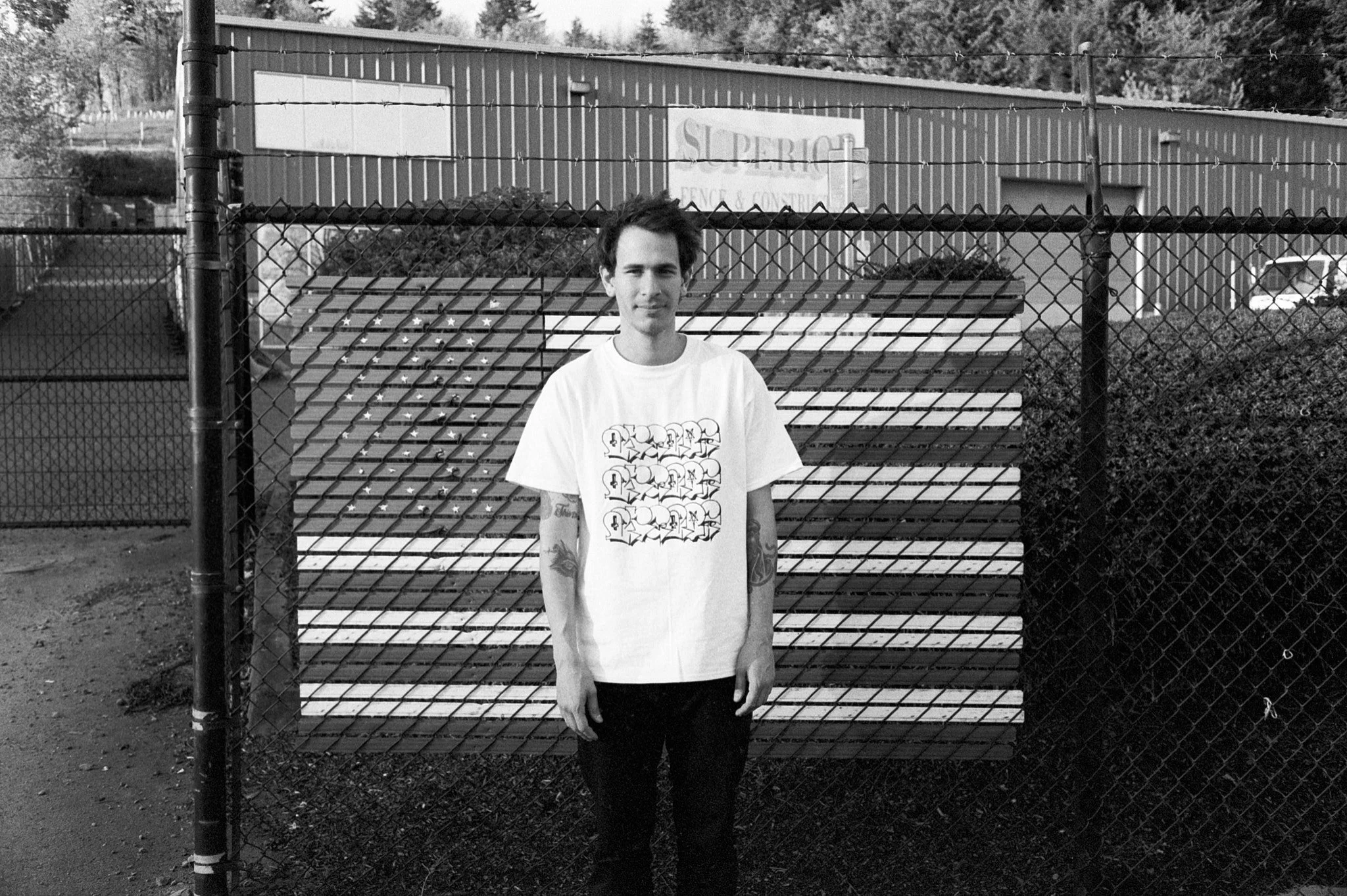 A young man with dark hair, wearing a white t-shirt with graphic print, standing in front of a chain-link fence and construction material, with a building and trees in the background.