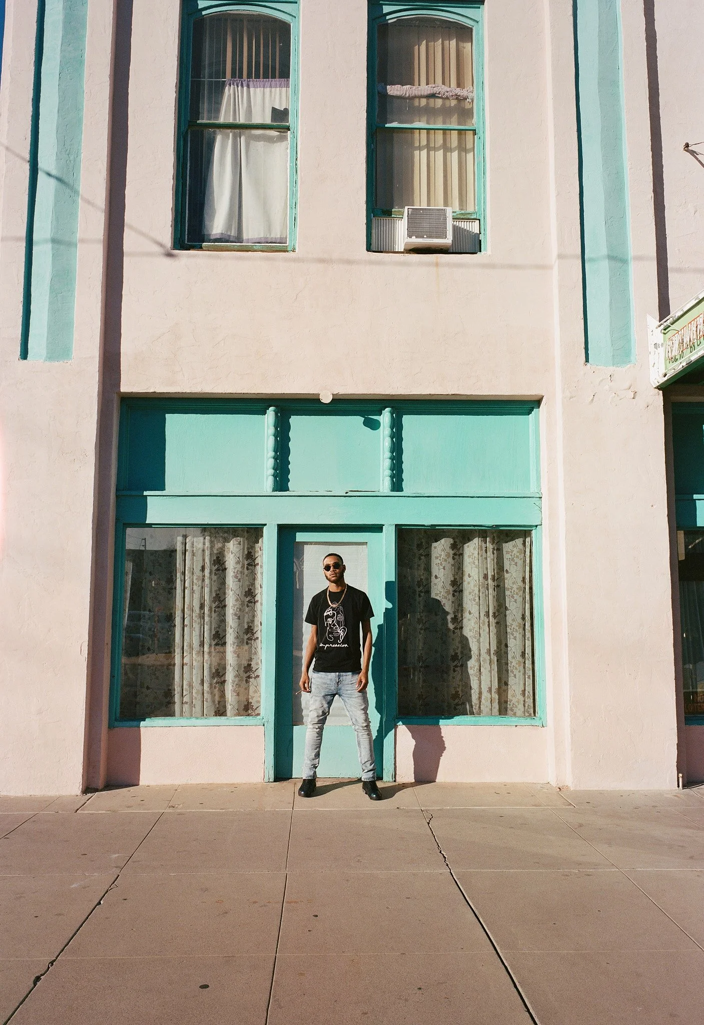 A man standing in front of a light pink building with turquoise window frames and door, wearing a black t-shirt, light jeans, black boots, sunglasses, and a gold chain.