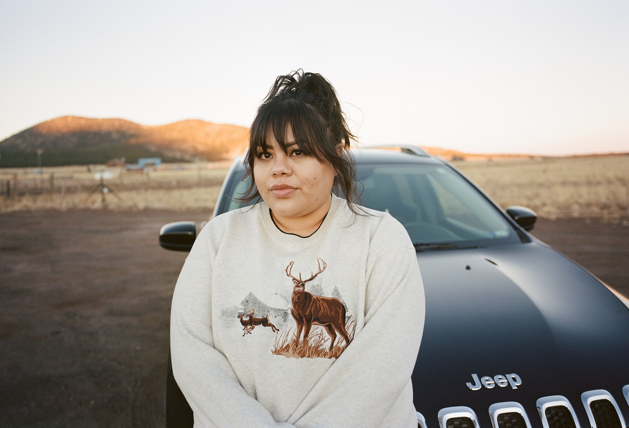Young woman with dark hair and bangs, wearing a light-colored sweatshirt with a deer graphic, standing in front of a black Jeep vehicle in an open landscape, with hills in the background during sunset.