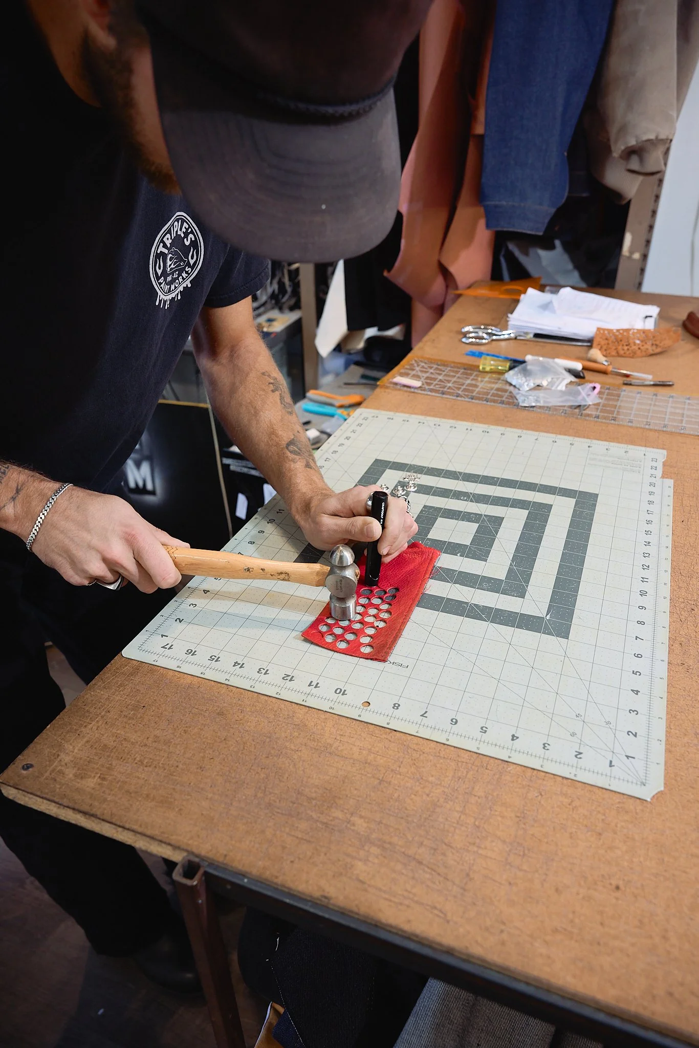 A person working on a craft project using a hammer and a steel punch on a red piece of material on a cutting mat.