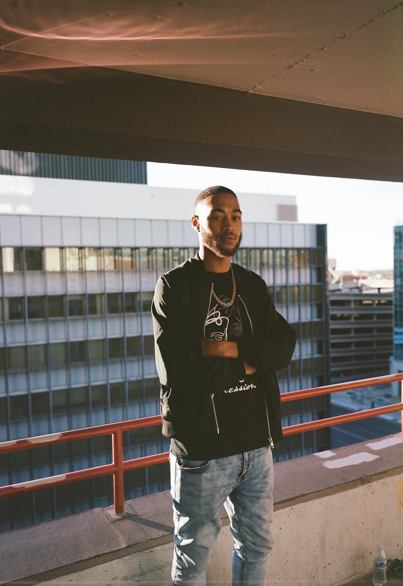 A young man stands on a rooftop parking garage with his arms crossed, wearing a black jacket, graphic T-shirt, light-wash jeans, gold chain, and is illuminated by natural sunlight with city buildings in the background.