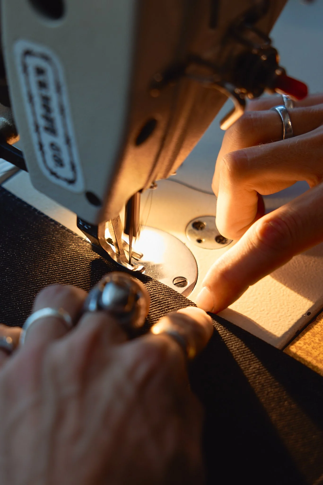 Close-up of hands guiding black fabric under a vintage sewing machine needle, with the machine illuminated by warm light.