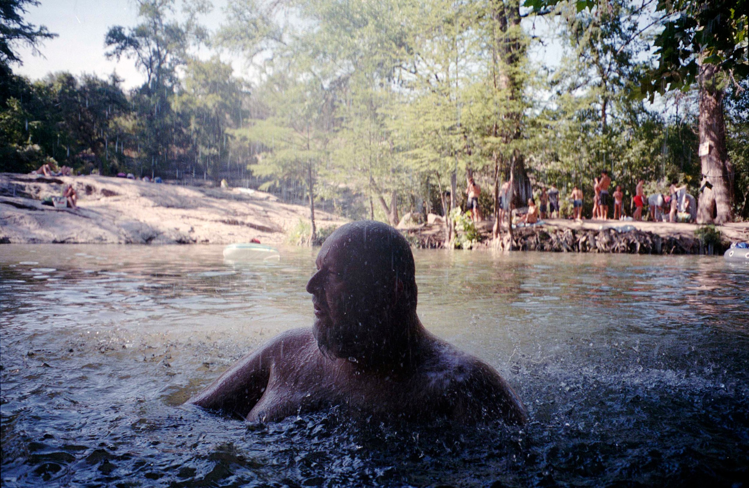 A person with a beard and short hair swimming in a river, with a group of people on the riverbank in the background, surrounded by trees.