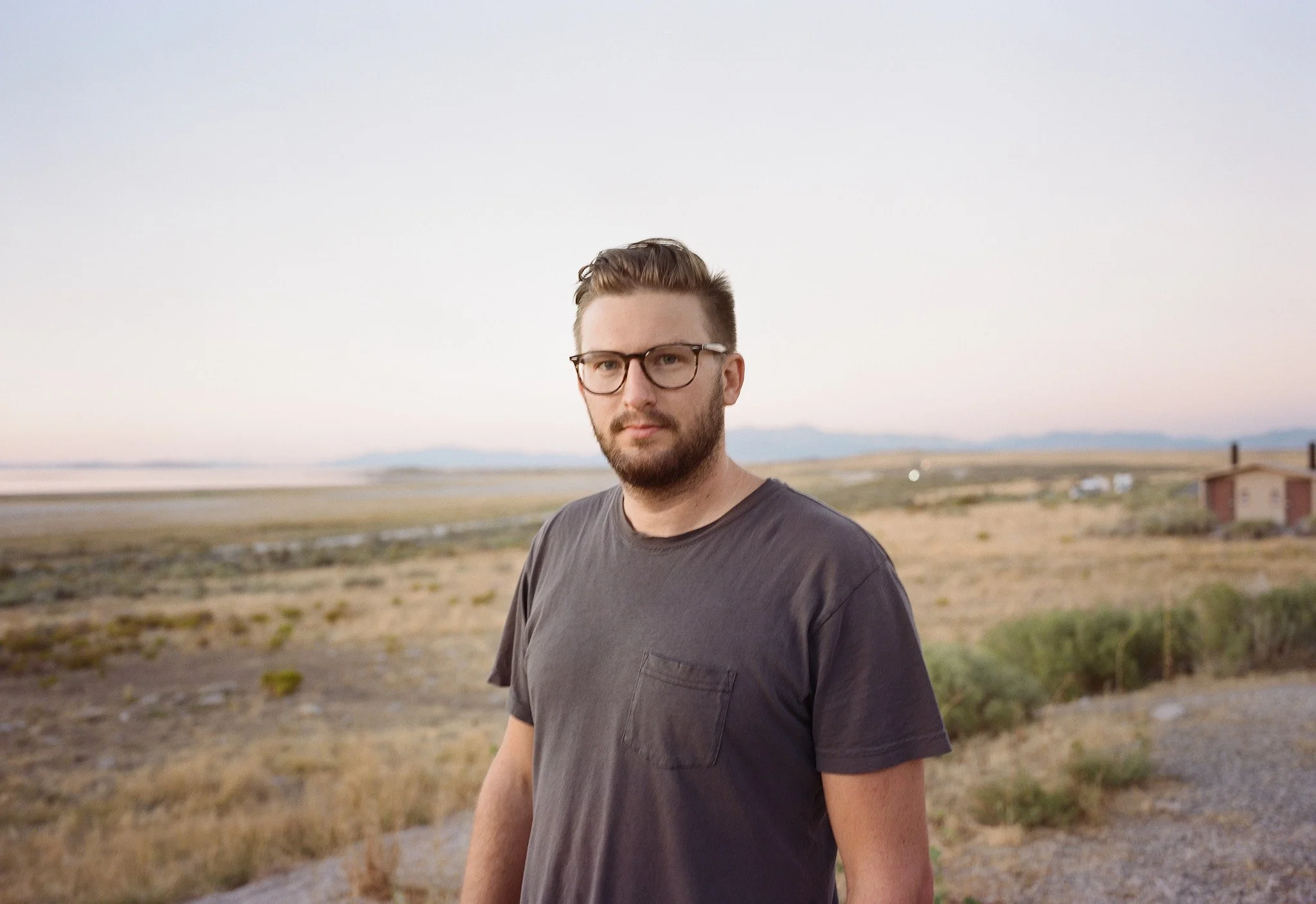 A young man with glasses and a beard stands outdoors in a dry, open landscape during dusk, looking into the camera.
