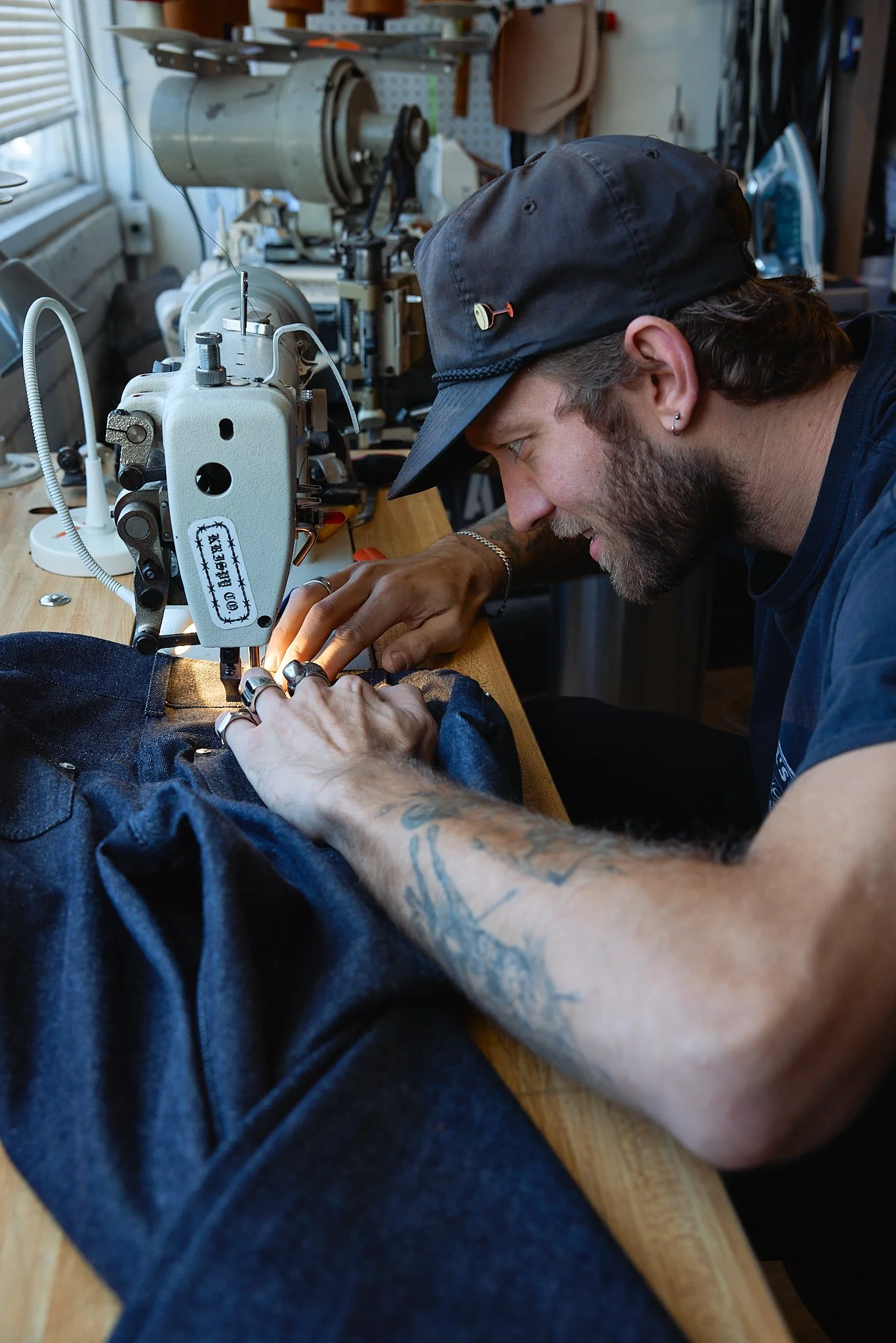 A man with tattoos sewing denim jeans on a vintage industrial sewing machine in a workshop.