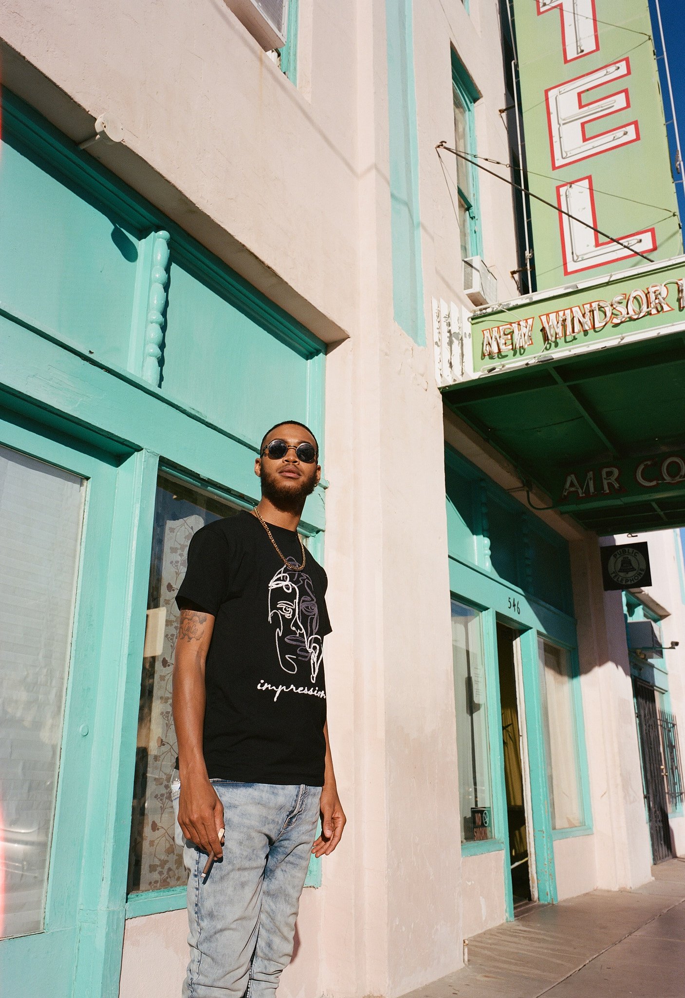 A man wearing sunglasses, a black graphic T-shirt, and gray jeans standing outside a colorful building with turquoise window frames. The building has a sign that reads 'NEW WINDSOR' and 'RELE', with part of the sign cut off. The man is holding a ciga