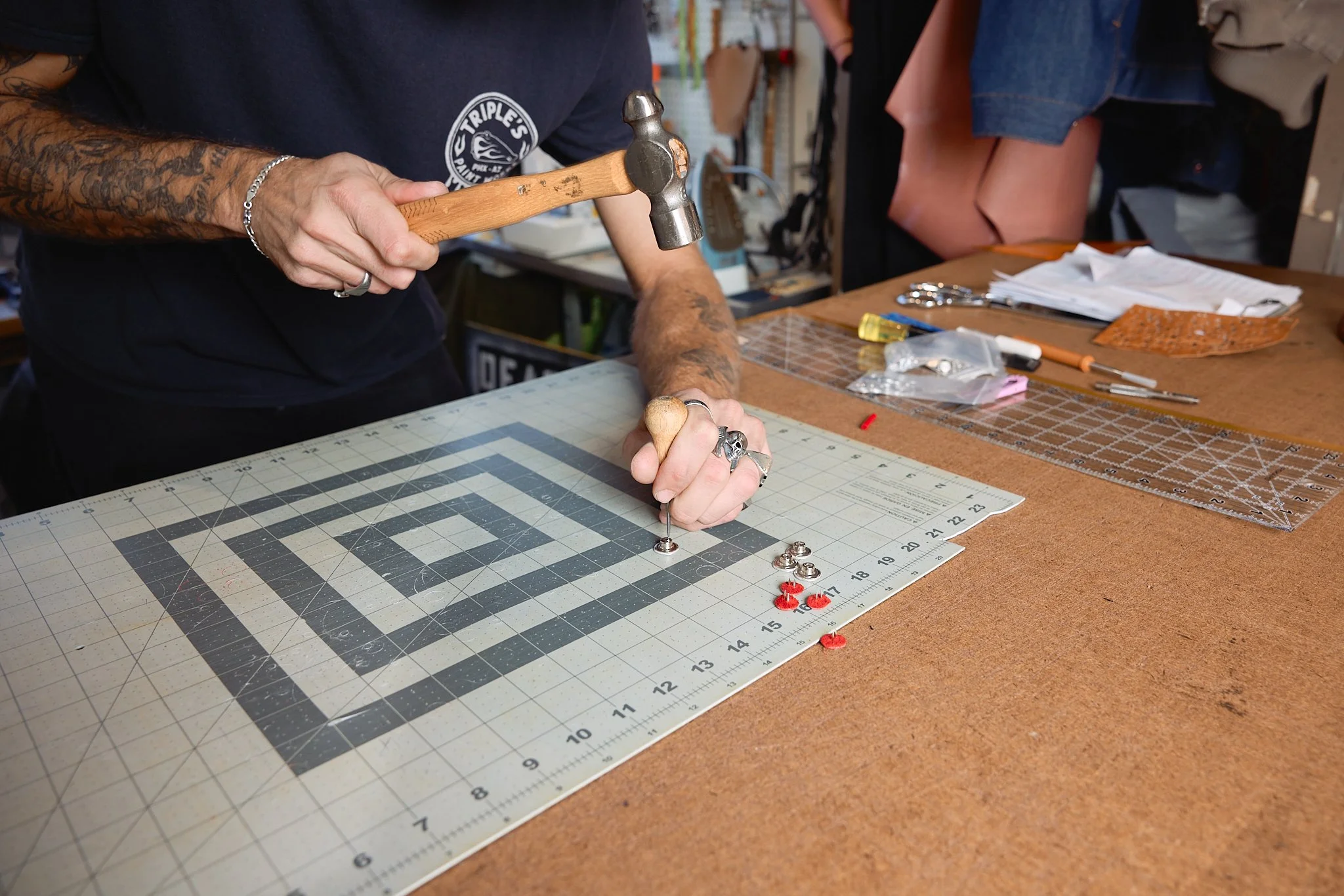 Person using a hammer and screwdriver to assemble jewelry on a workbench with various tools and papers around.