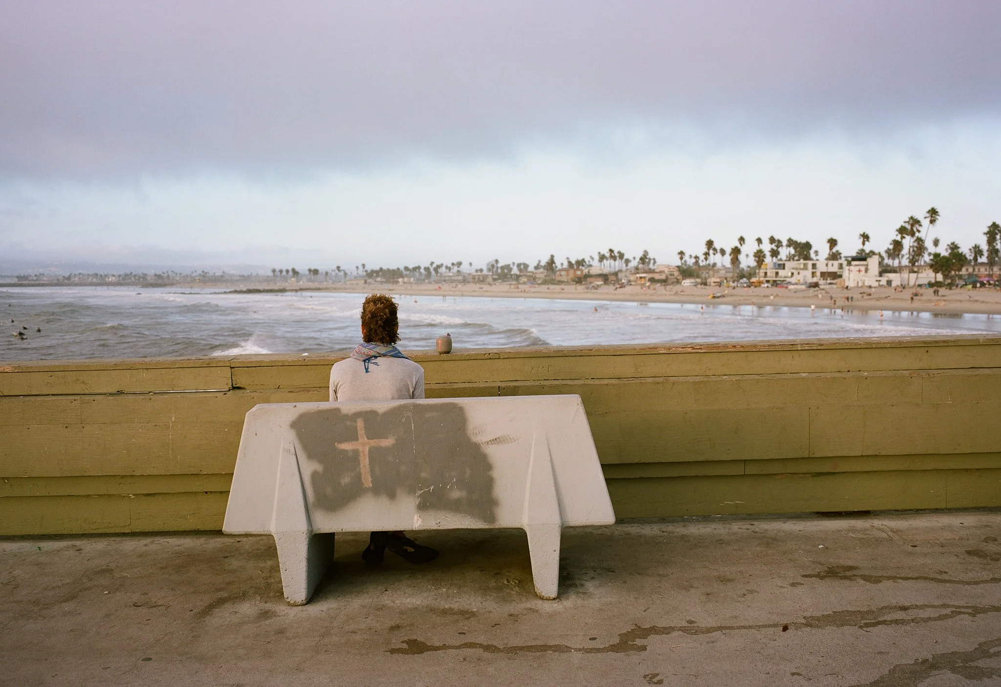 A person sitting on a bench at a beach pier, facing the ocean with houses and palm trees in the background, overcast sky.