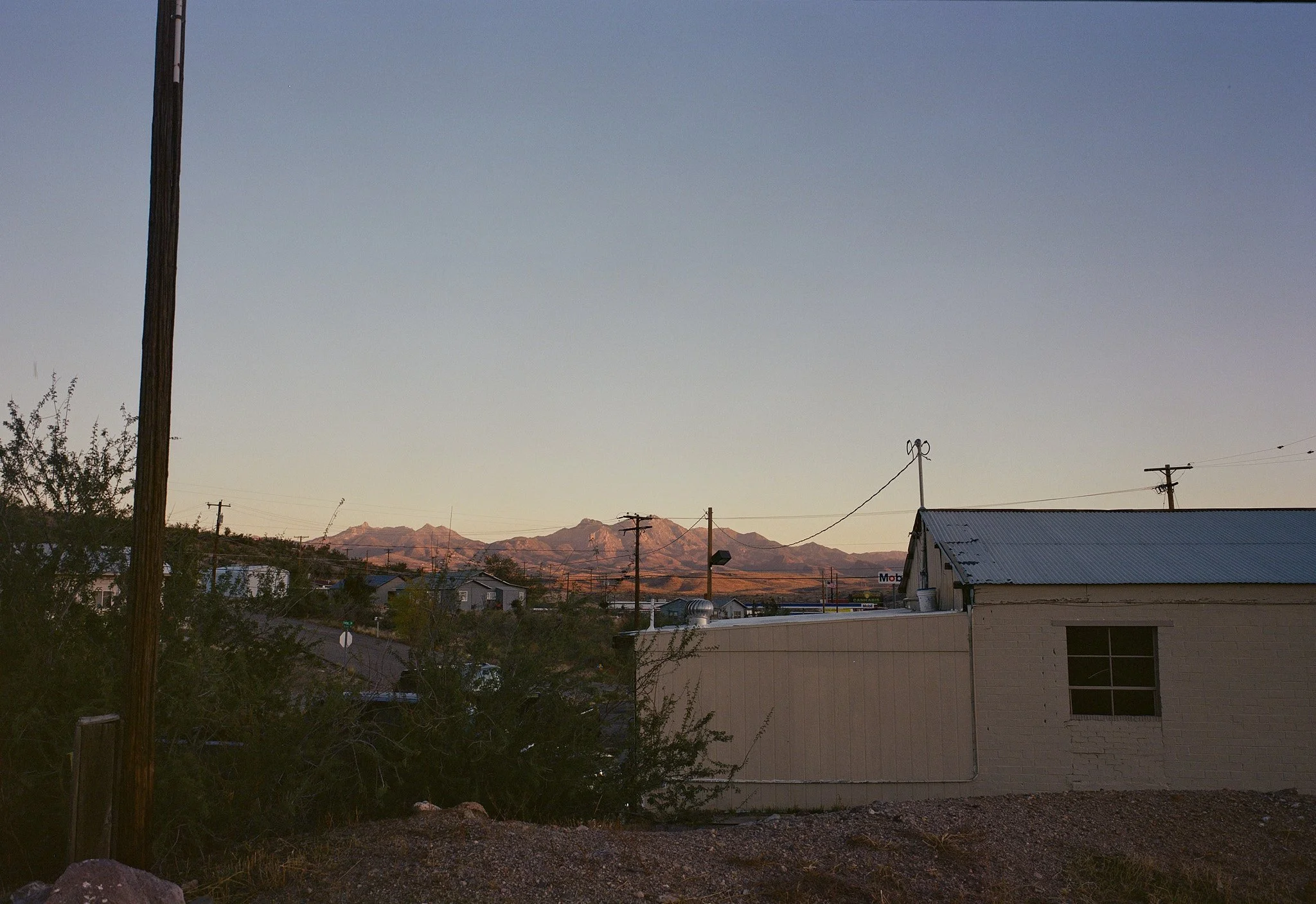 A rural landscape at sunset with mountains in the background, utility poles, and a small building with a metal roof in the foreground.
