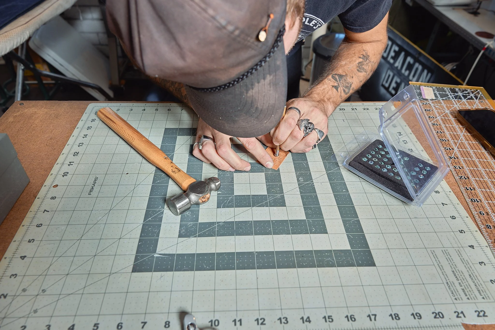 Person working at a crafting or jewelry making station, using a small tool and a ruler on a cutting mat. Accessories include rings and tattoos on both arms. Items on the table include a hammer, a small plastic container with screws, and a smartphone.